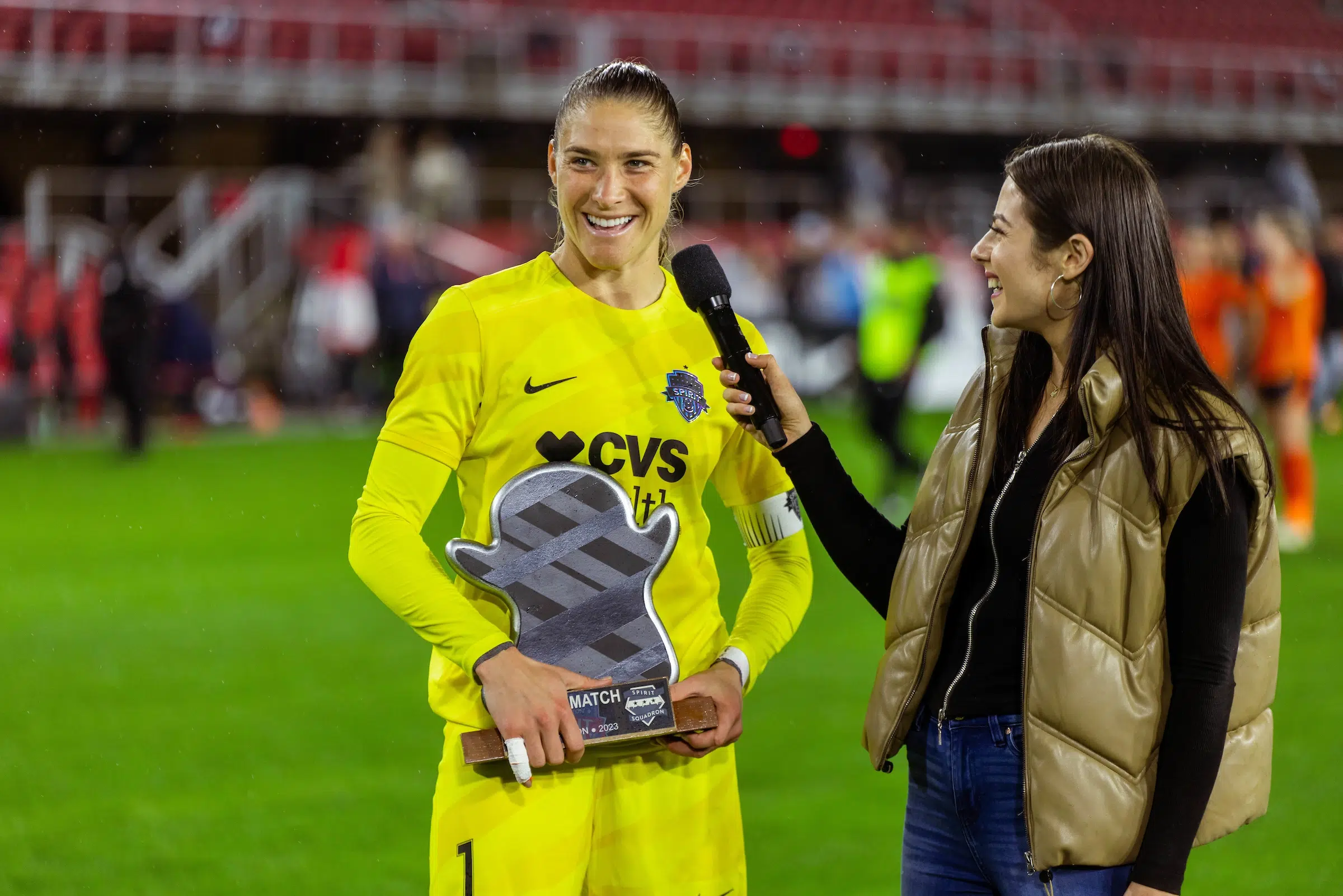 Aubrey Kingsbury in a yellow uniform holds a ghost-emoji shaped trophy while she is interviewed by a woman in a black long sleeve top and tan leather vest.