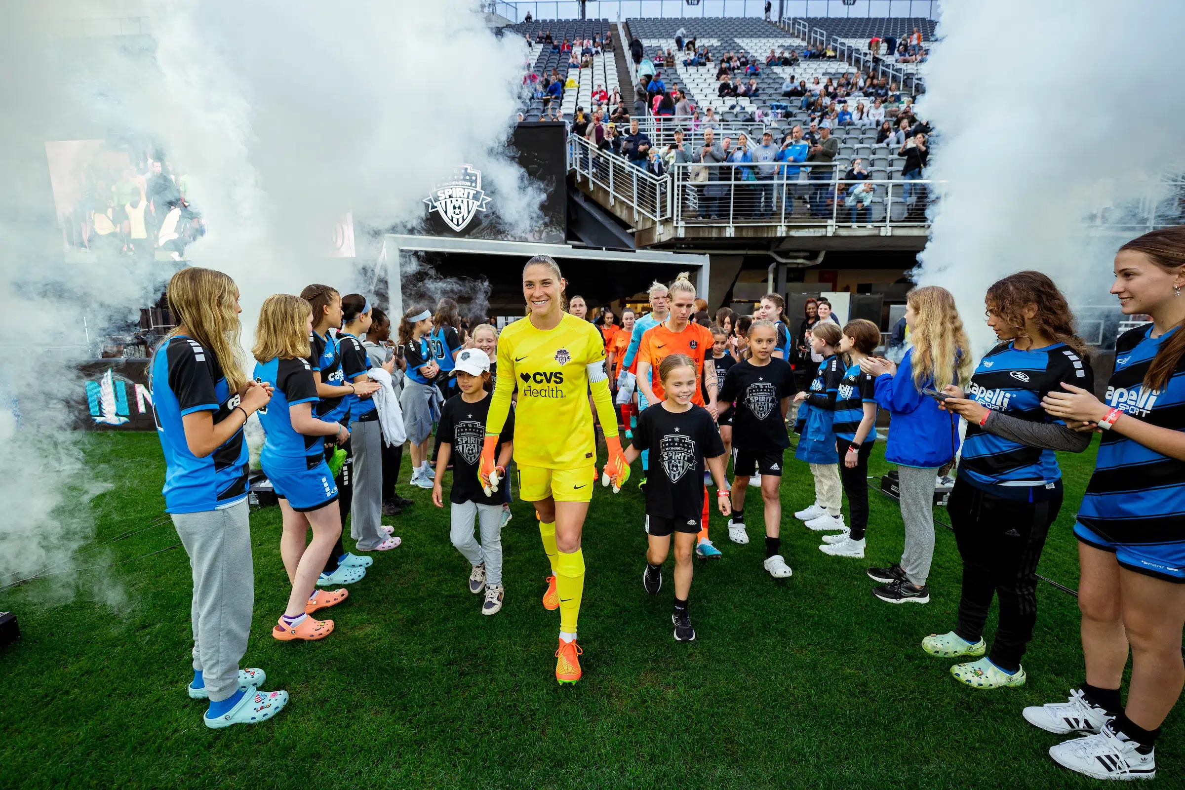 Aubrey Kingsbury in a yellow goalie uniform walks out of a tunnel holding hands with two young girls in black tshirts. Behind her, two teams of players walk out of the tunnel holding hands with children. A row of preteen girls lines both sides of the players as smoke is shot into the sky.