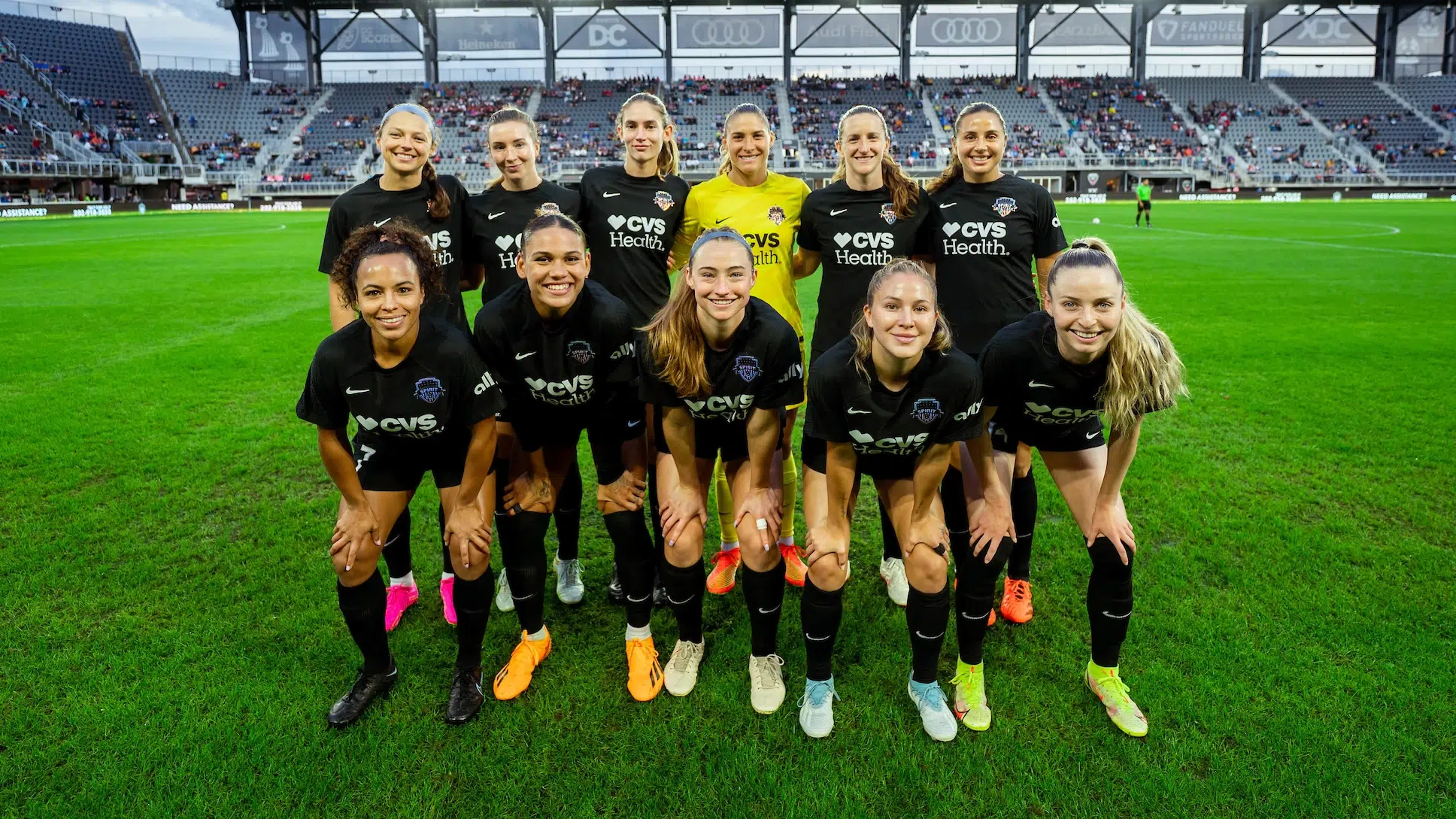 10 soccer players in all black uniforms and one in a yellow goalie uniform pose in two rows for a photo on a soccer field with bleachers in the background.