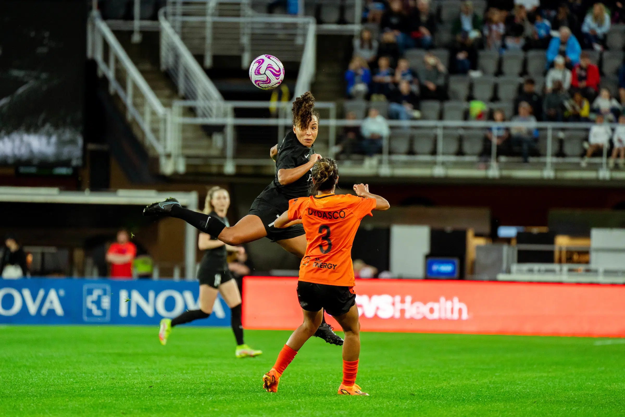 Ines Jaurena leaps in the air to head a soccer ball. A defender in an orange uniform remains on the ground.