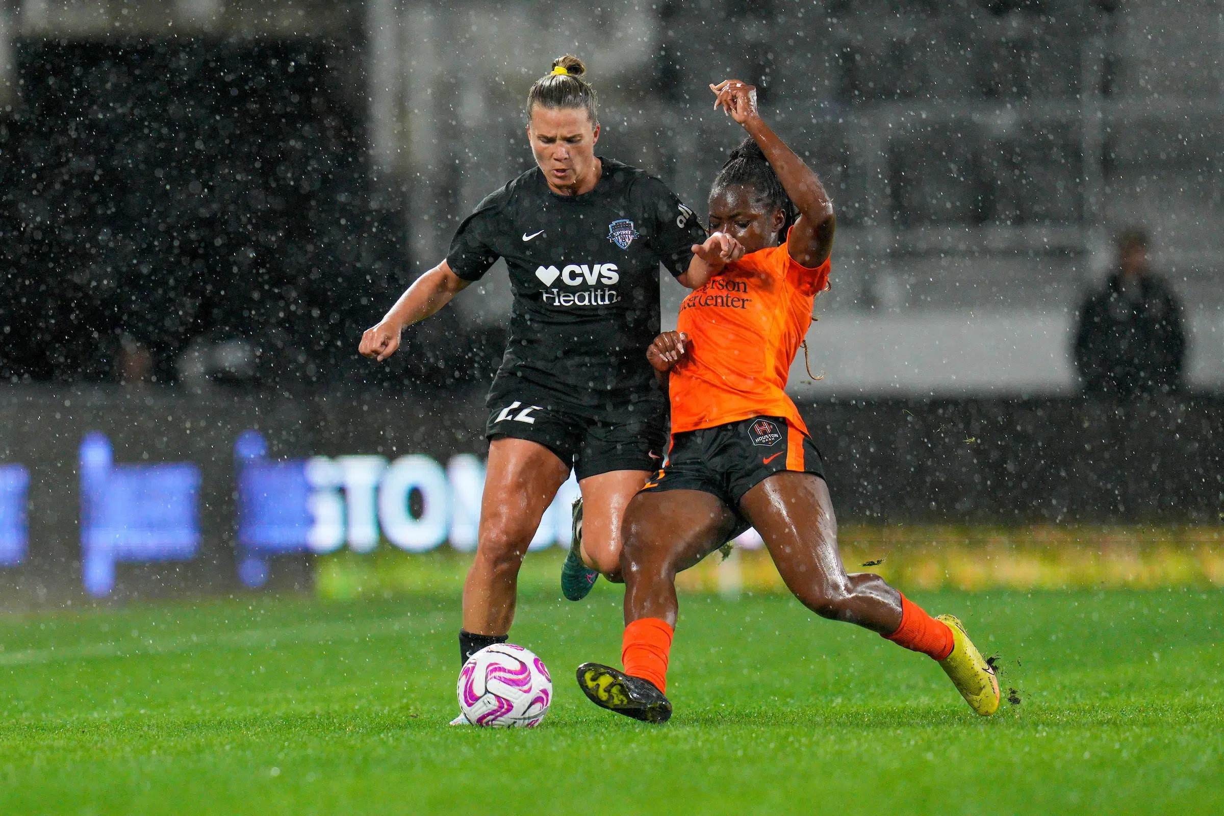 Amber Books in a black uniform dribbles a ball as a defender in an orange uniform attempts to tackle the ball away from her. It's raining.