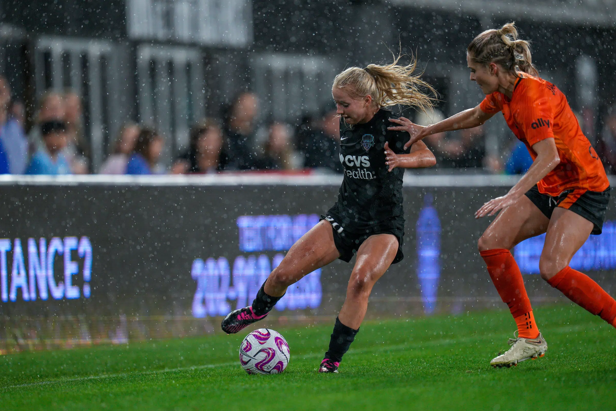 Chloe Ricketts in a black uniform works to keep a soccer ball away from a defender in an orange uniform as rain pours down.