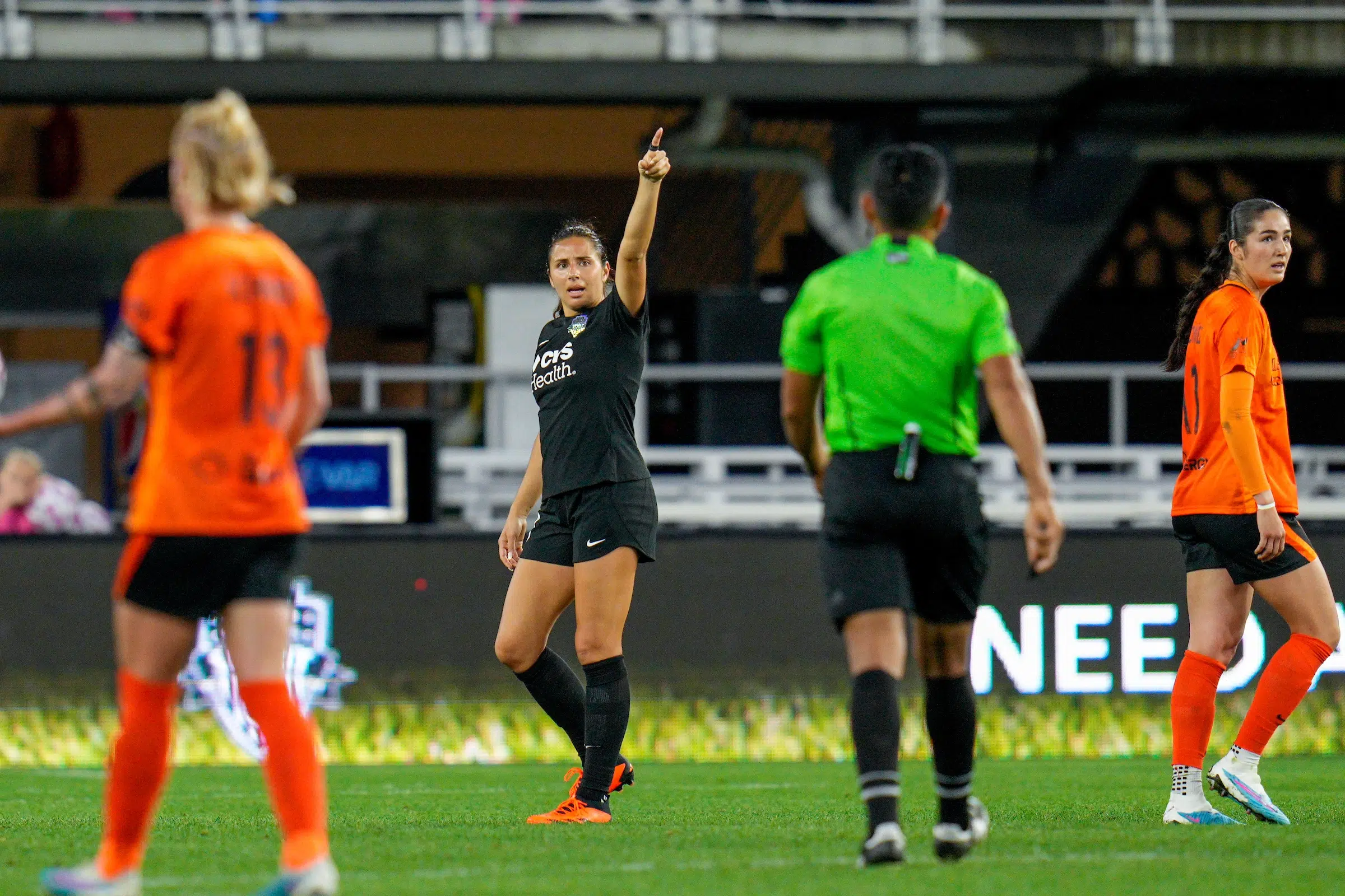 Sam Staab in an all black uniform points at a referee in a green shirt on a soccer field.