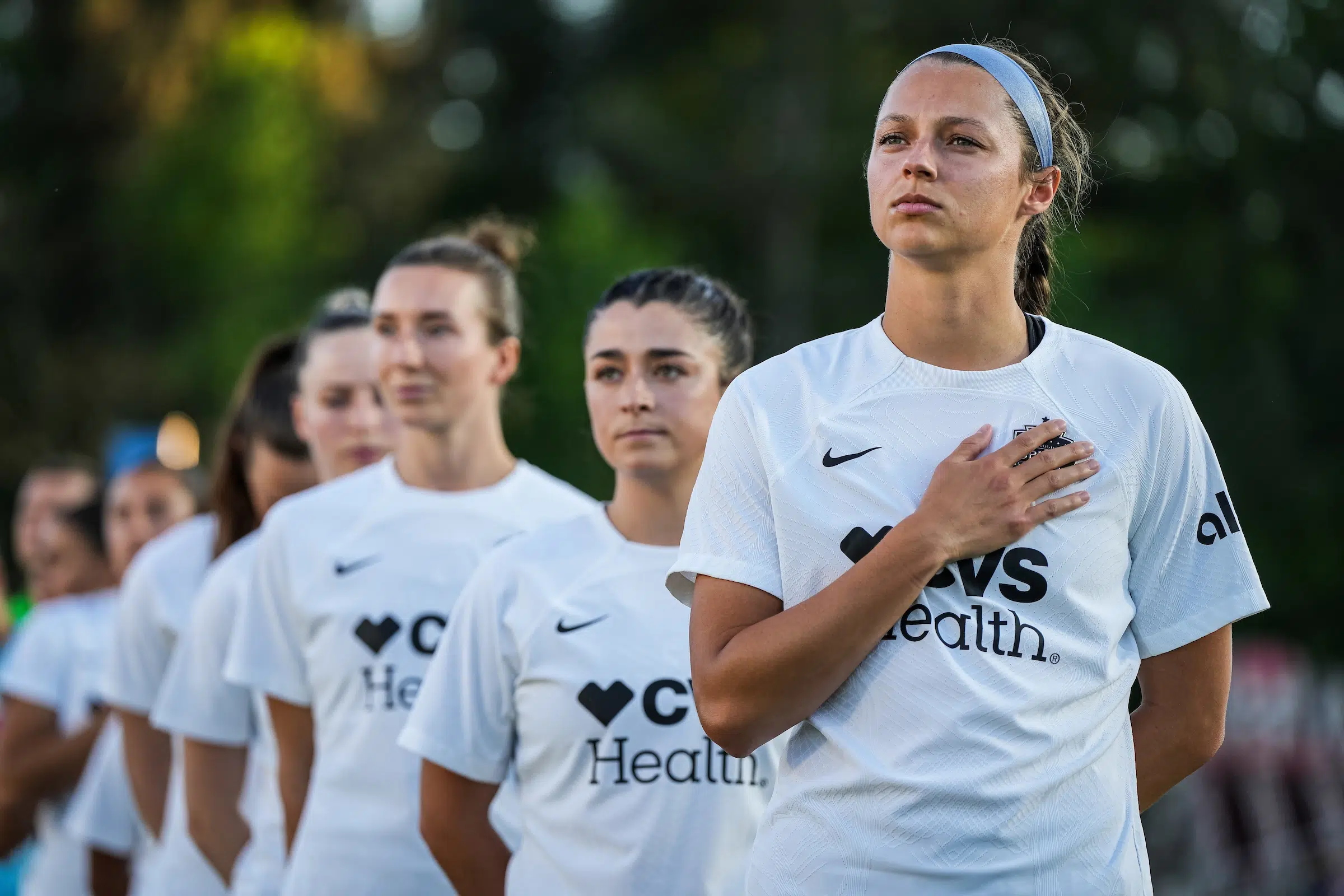 Ashley Hatch in a white uniforms stands at the front of a line of players in matching uniforms. She has her hand on her heart and looks off into the distance.