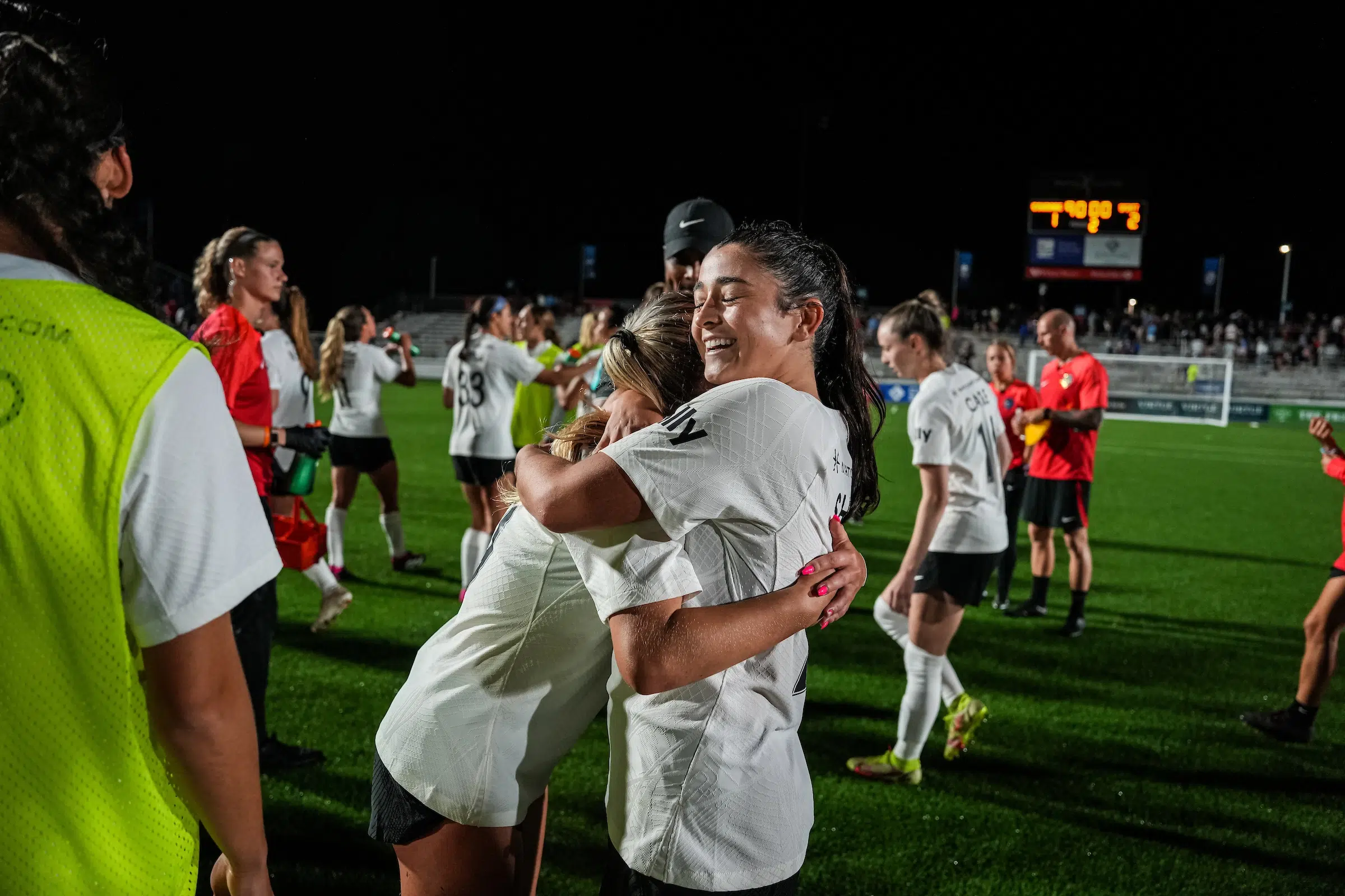 Two soccer players in white tops and black shorts embrace and smile. In the background, an out-of-focus scoreboard shows the final score of 2 - 1.