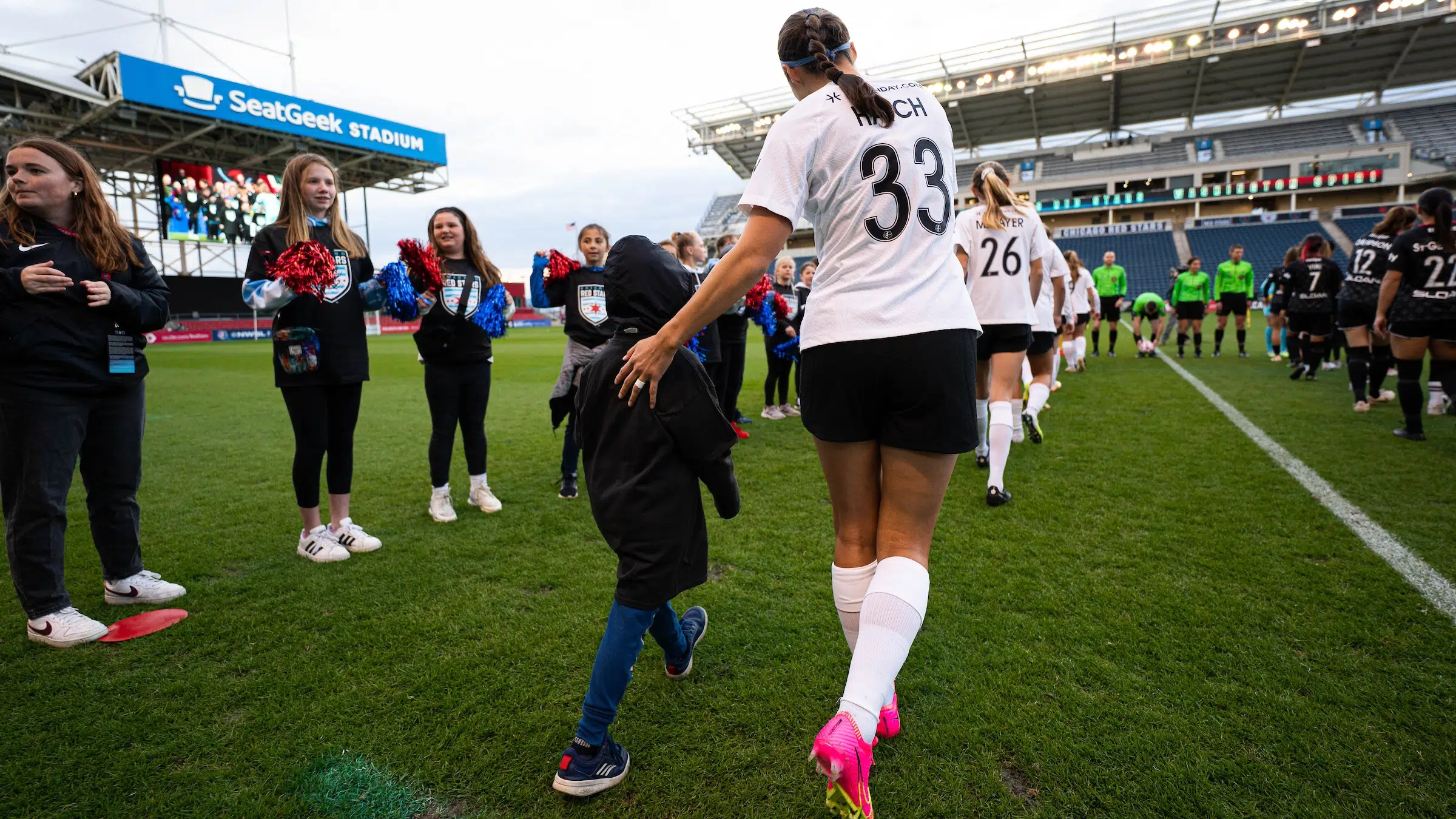 Ashley Hatch in a white top, black shorts, and white socks leads a young fan out onto the soccer field.