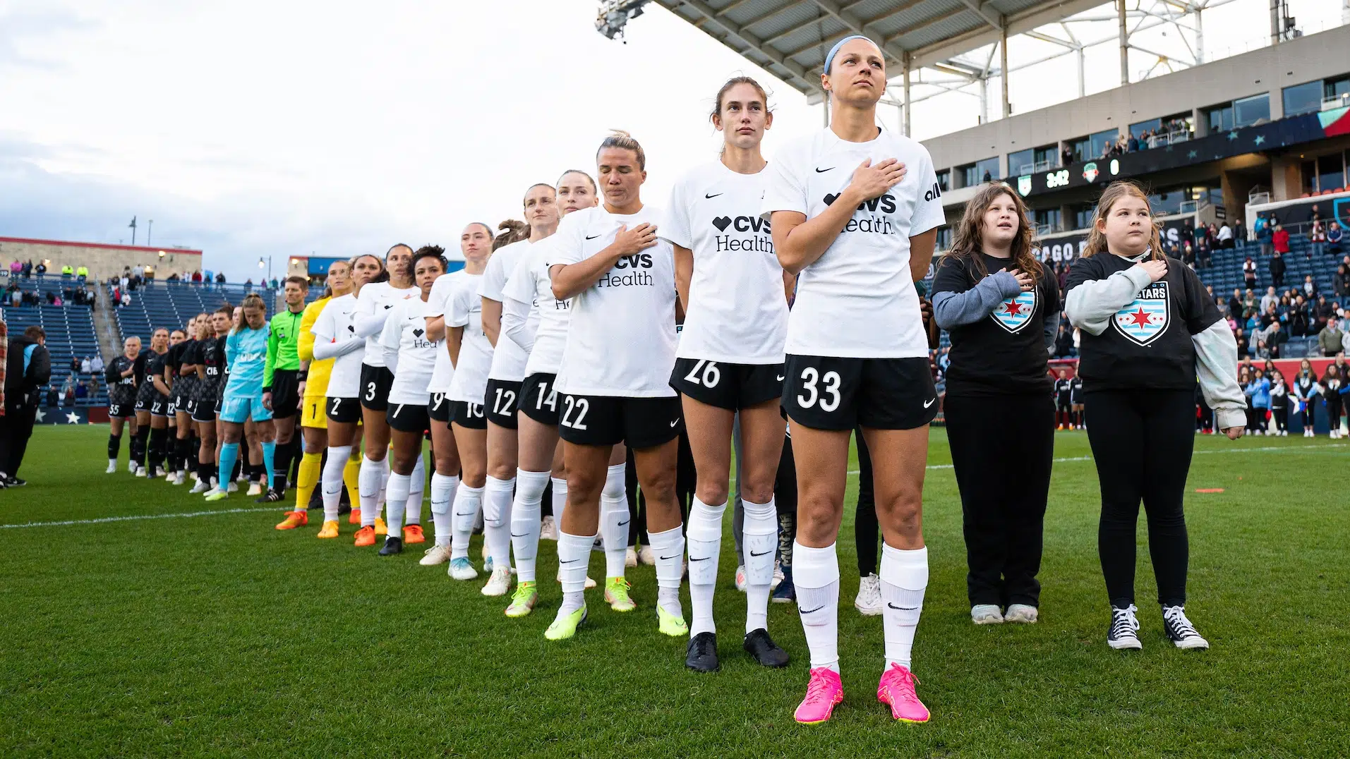 A team of soccer players in white tops, black shorts and white socks stand stoically in a line with their hands on their hearts.