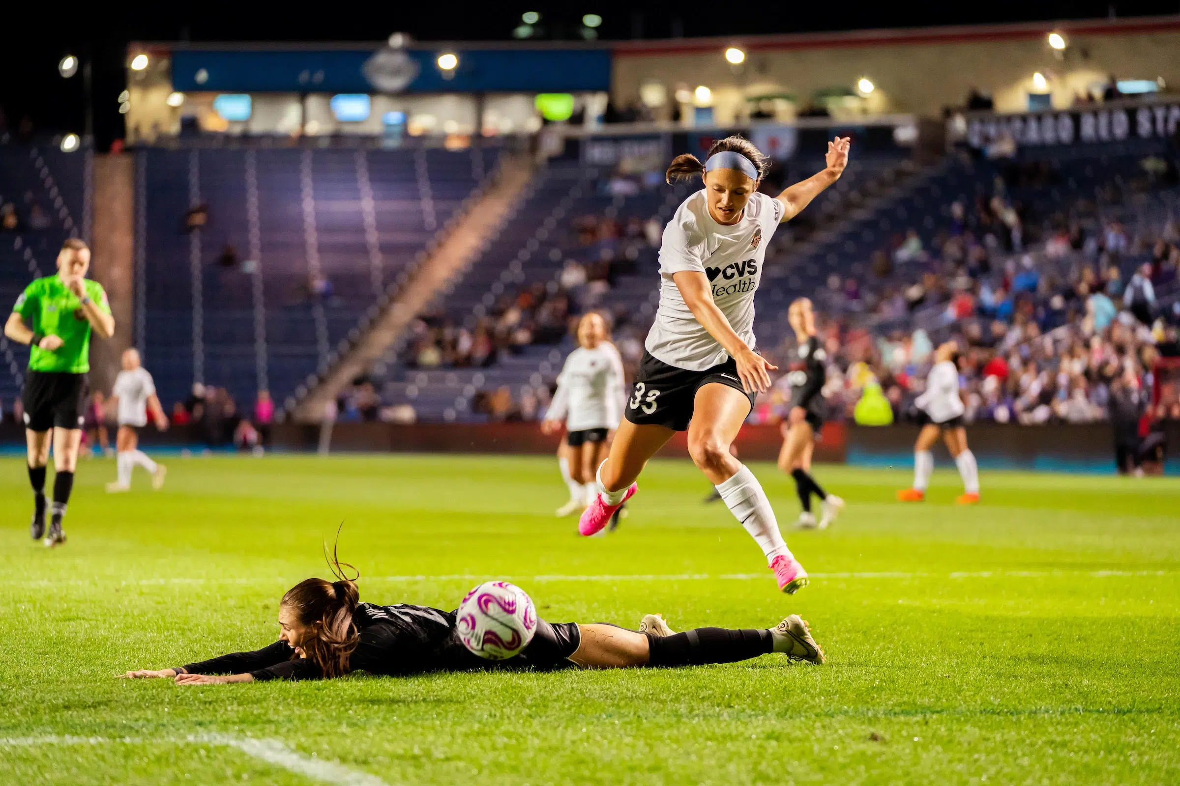 Ashley Hatch in a white top, black shorts, white socks and pink cleats jumps over a defender in a black uniform who lies on the ground.