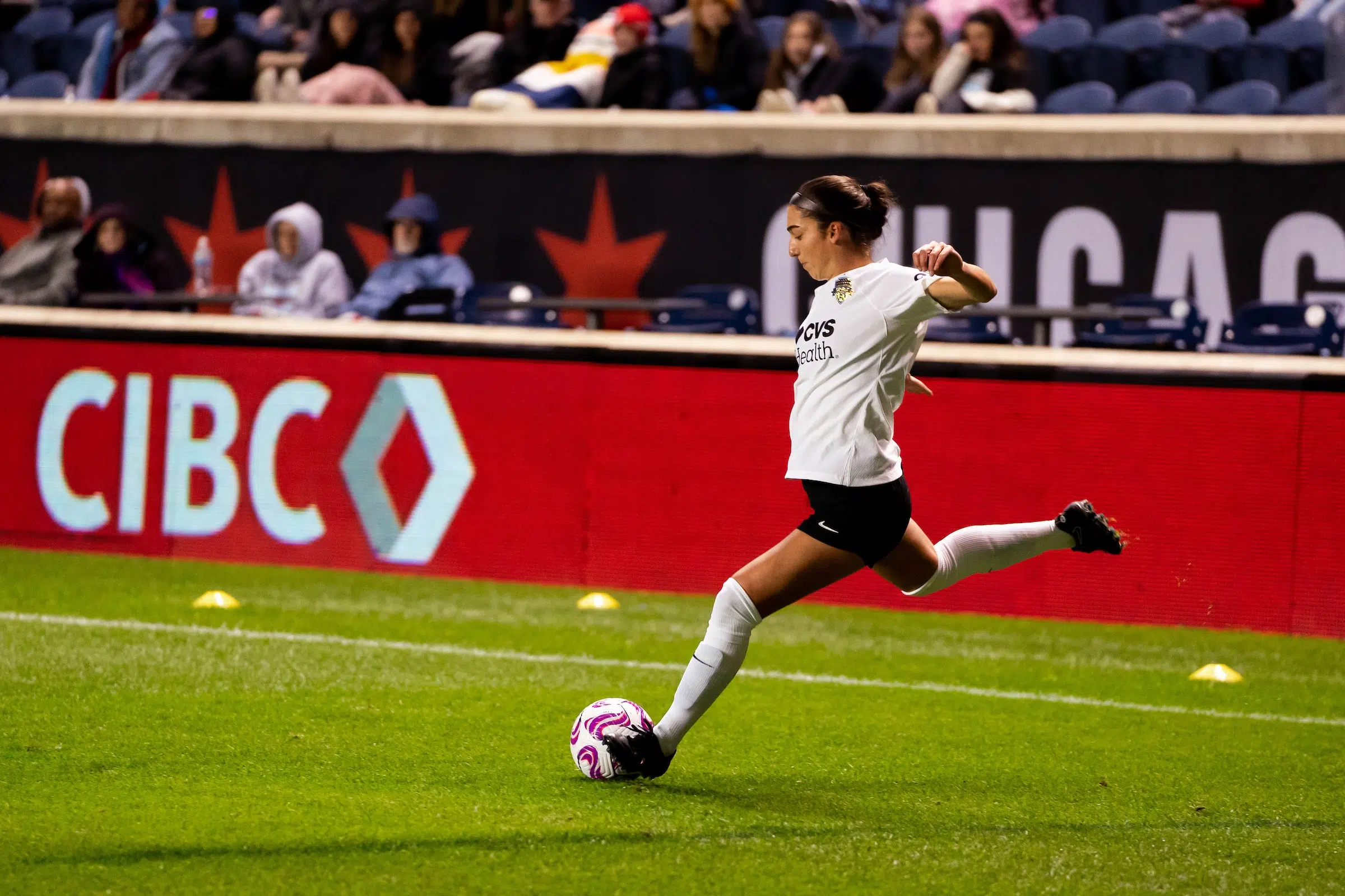 Lena Silano in a white top, black shorts and white socks winds up to cross a soccer ball towards goal.
