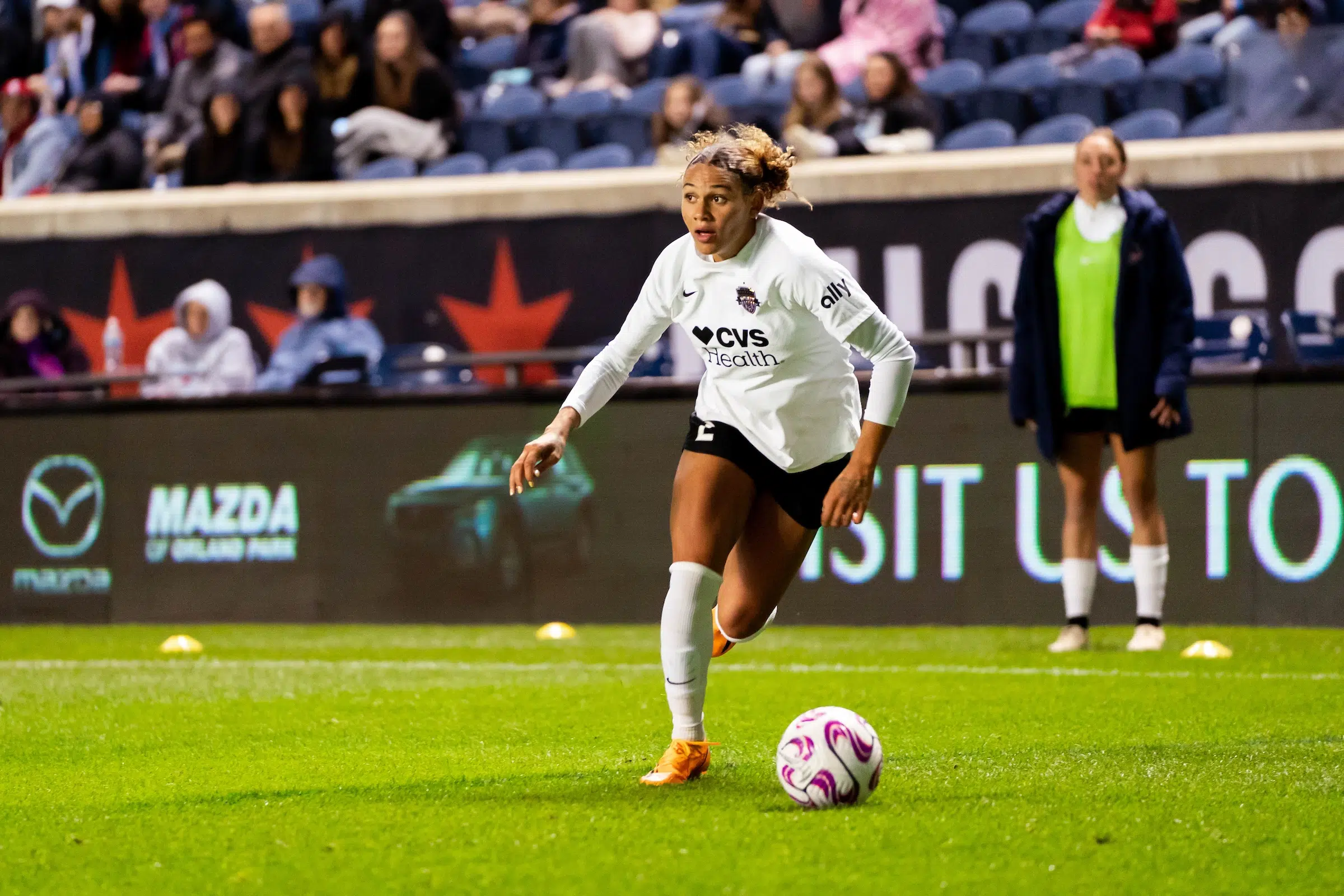 Trinity Rodman looks up to find her teammates as she dribbles a soccer ball. She wears a long sleeve white top, black shorts, and white socks.