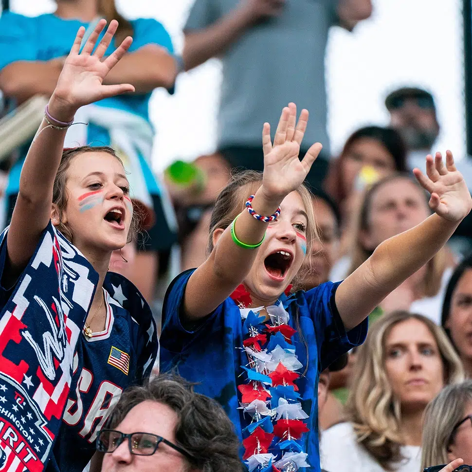 Two young fans in Washington Spirit and USWNT merch waves their hands and cheer.