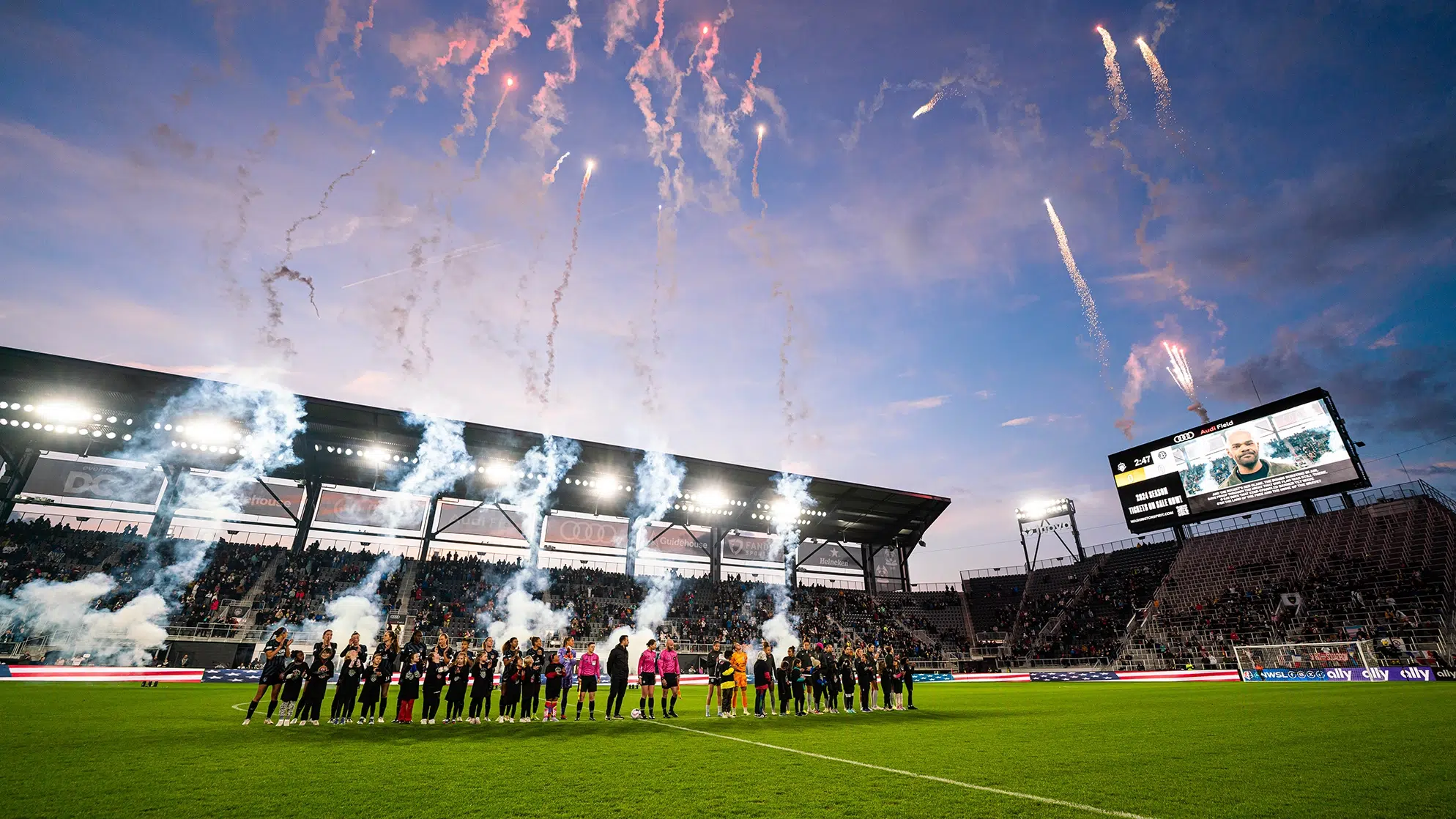 Soccer players stand at midfield of Audi Field while fire works are launched into the air.