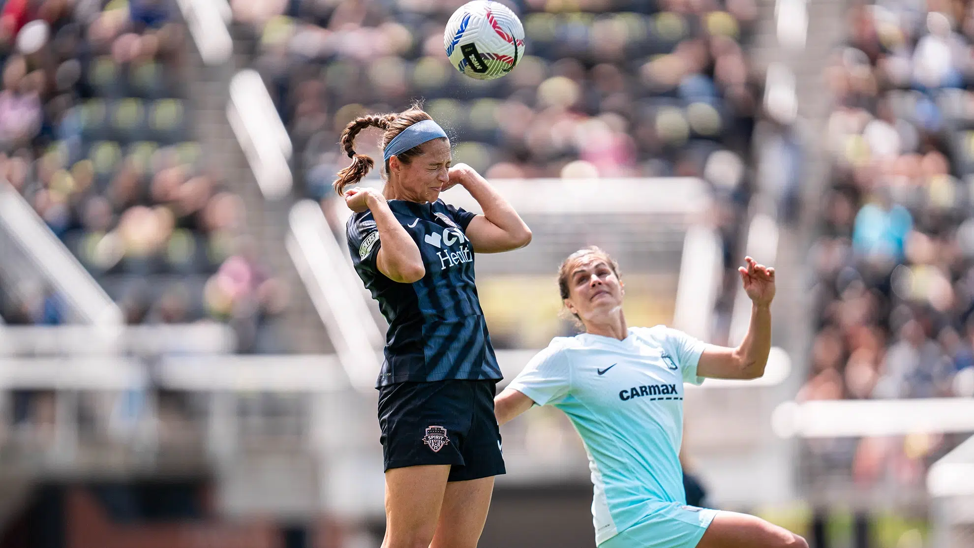 Ashley Hatch jumps up to head a soccer ball while a Gotham defender is seen below. Ashley Hatch is wearing a black Spirit kit with her hair in a braid.