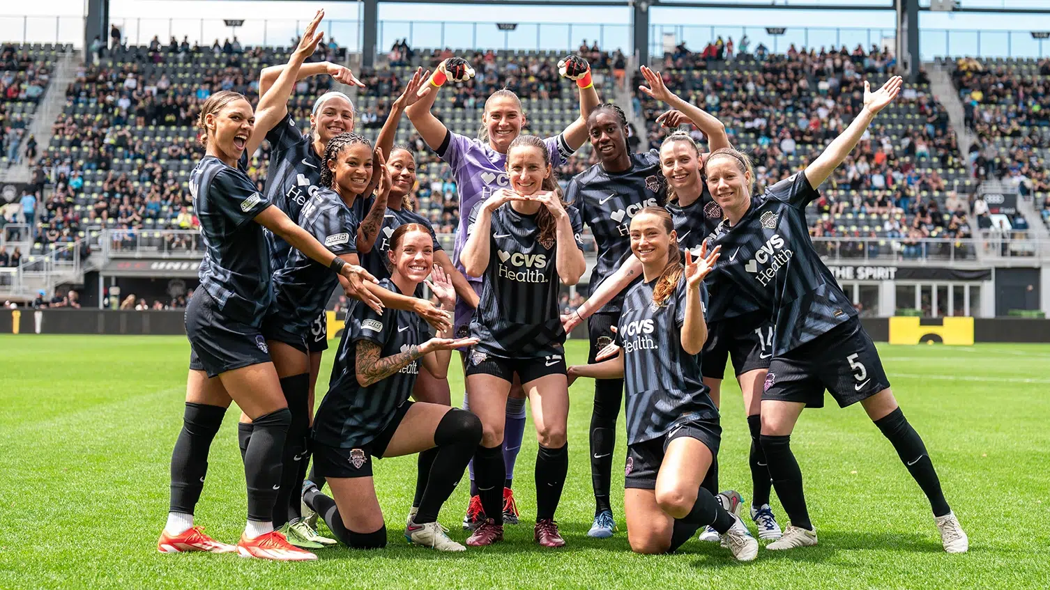 The starting eleven of the Washington Spirit pose for a photo with Andi Sullivan in the middle. All the other plays point towards her. They all are wearing black Spirit kits.