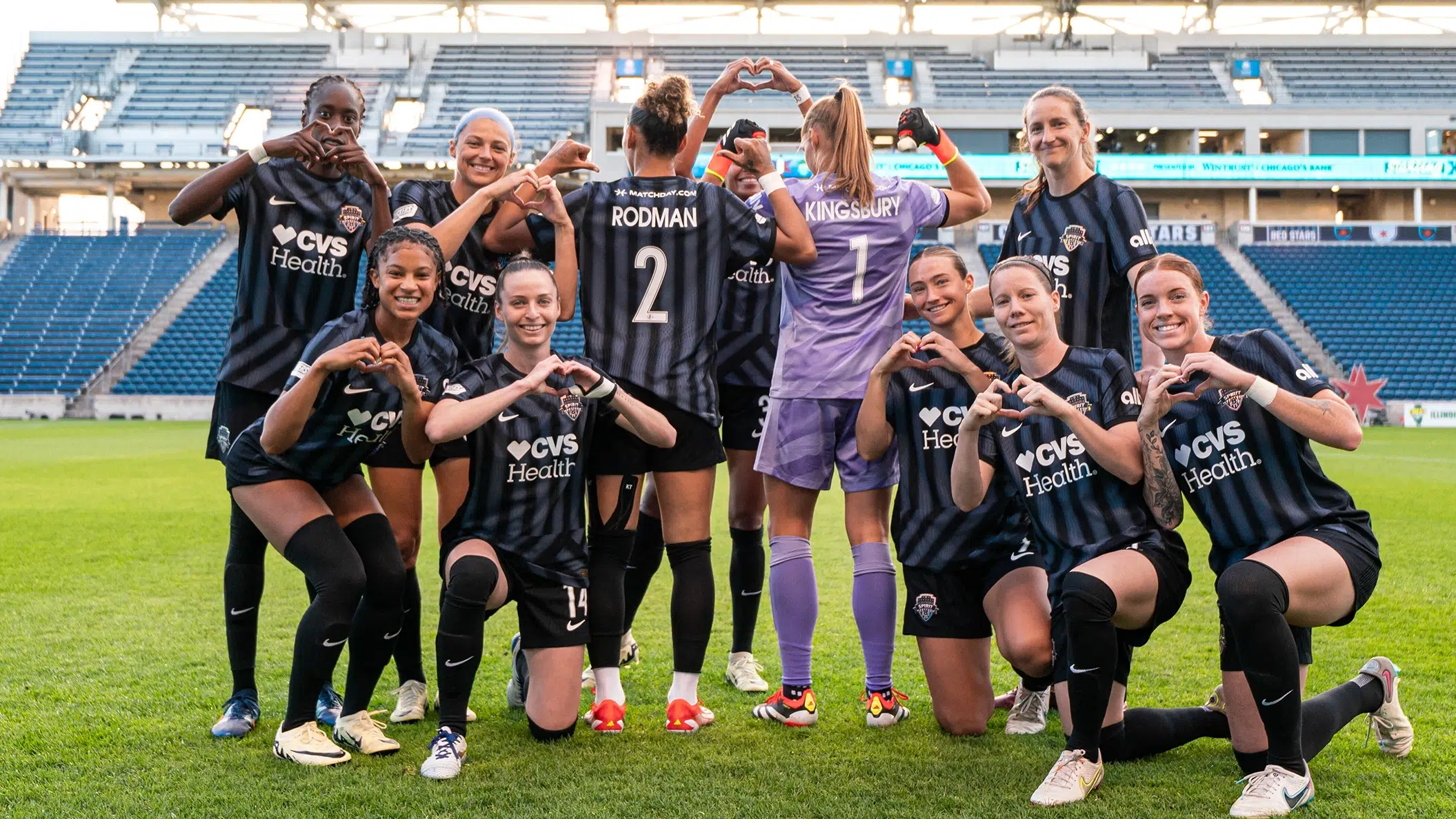 The starting XI of the Spirit hold up heart shapes with their hands as Trinity Rodman #2 and Aubrey Kingsbury #1 face away from the camera in the center.