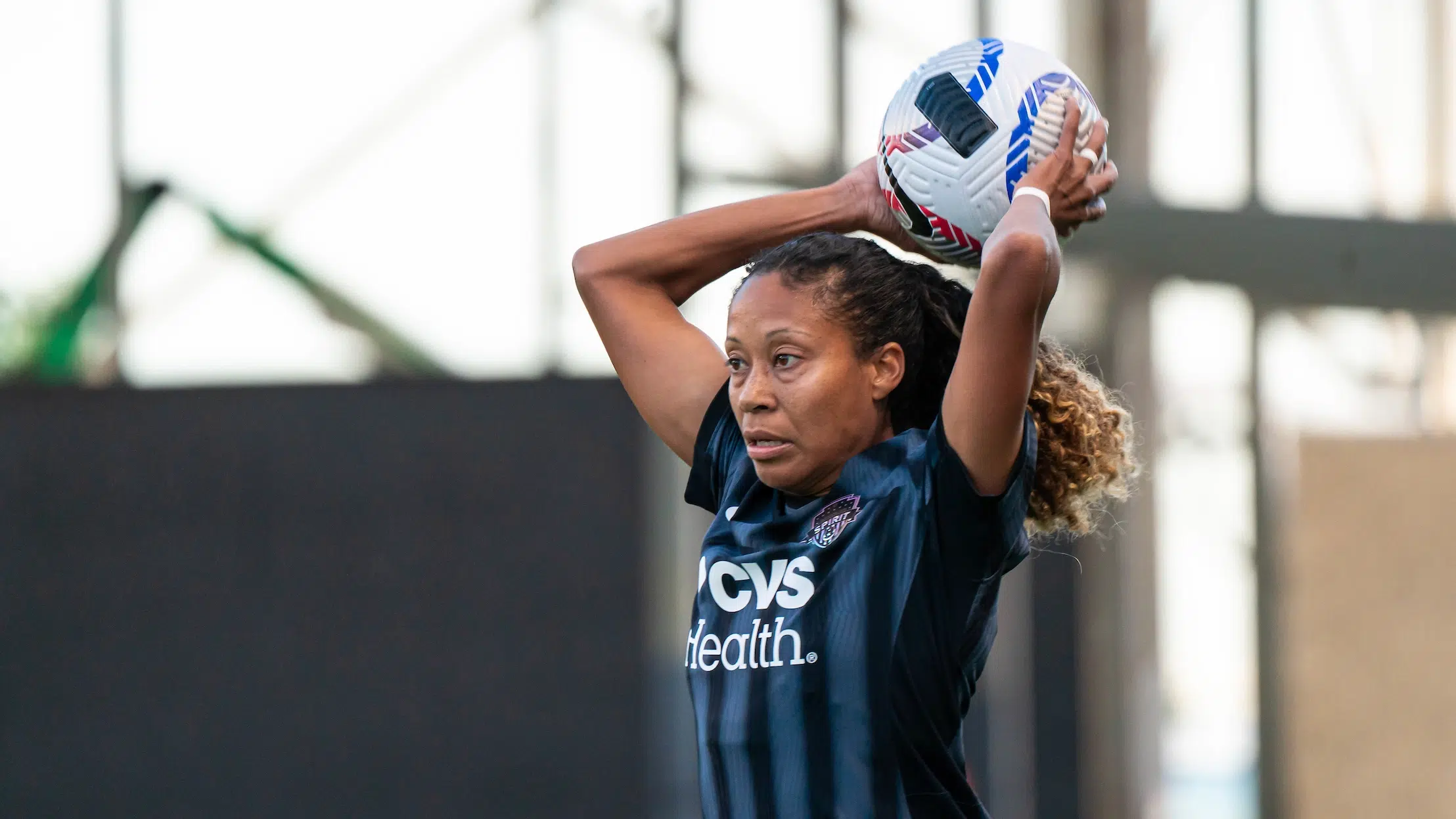 Casey Krueger in a black and gray striped jersey holds the soccer ball behind her head and prepares to throw.
