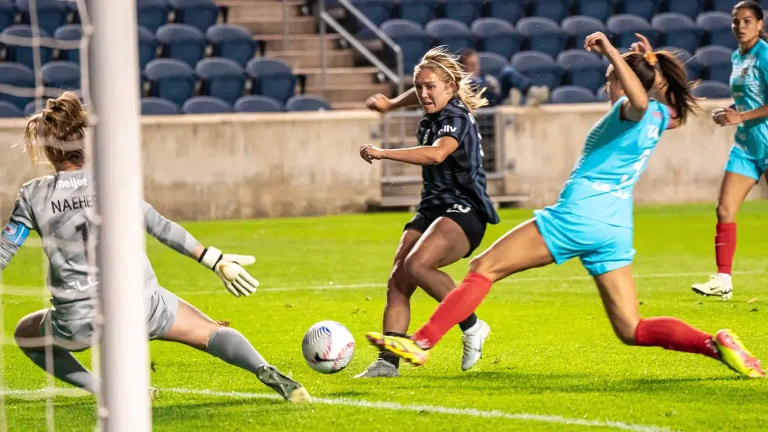 Brittany Ratcliffe slips the ball between the goalkeeper and a defender.