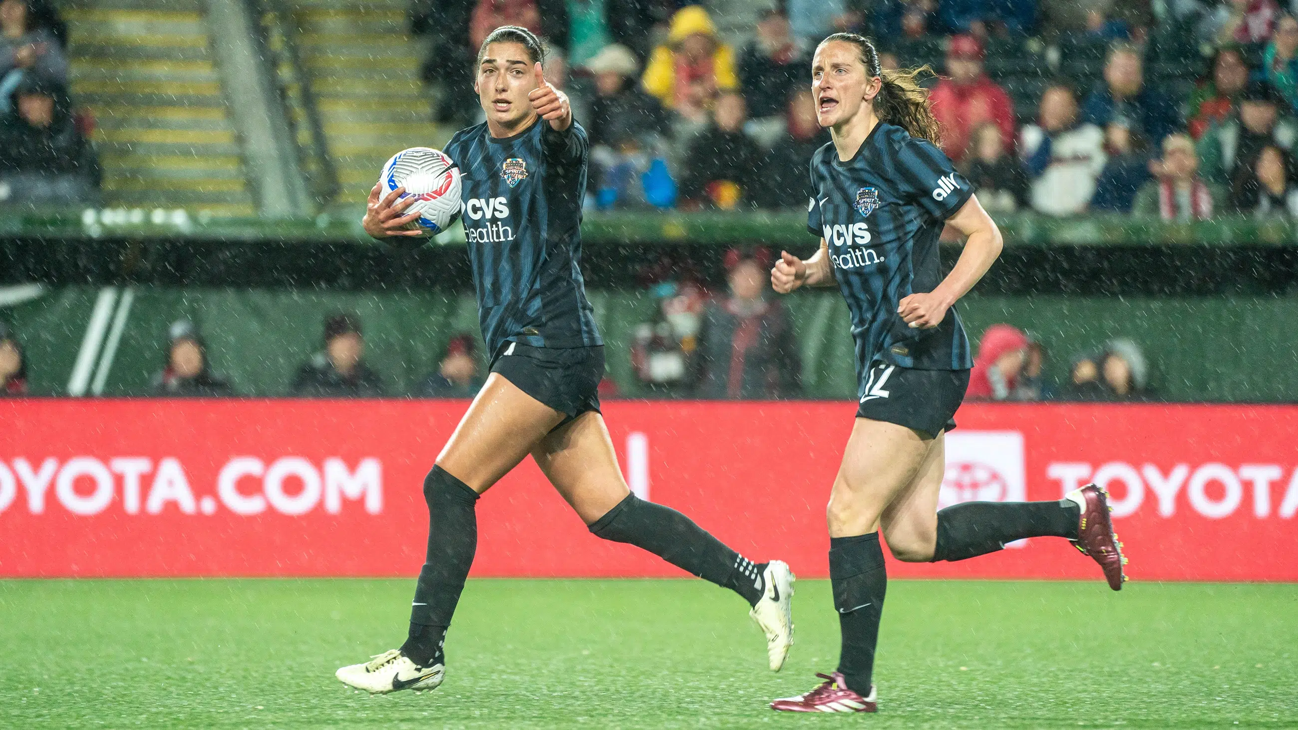 Lena Silano holds the soccer ball with one hand and gives a thumbs up with the other. Andi Sullivan runs besides her. Both are wearing black kits.