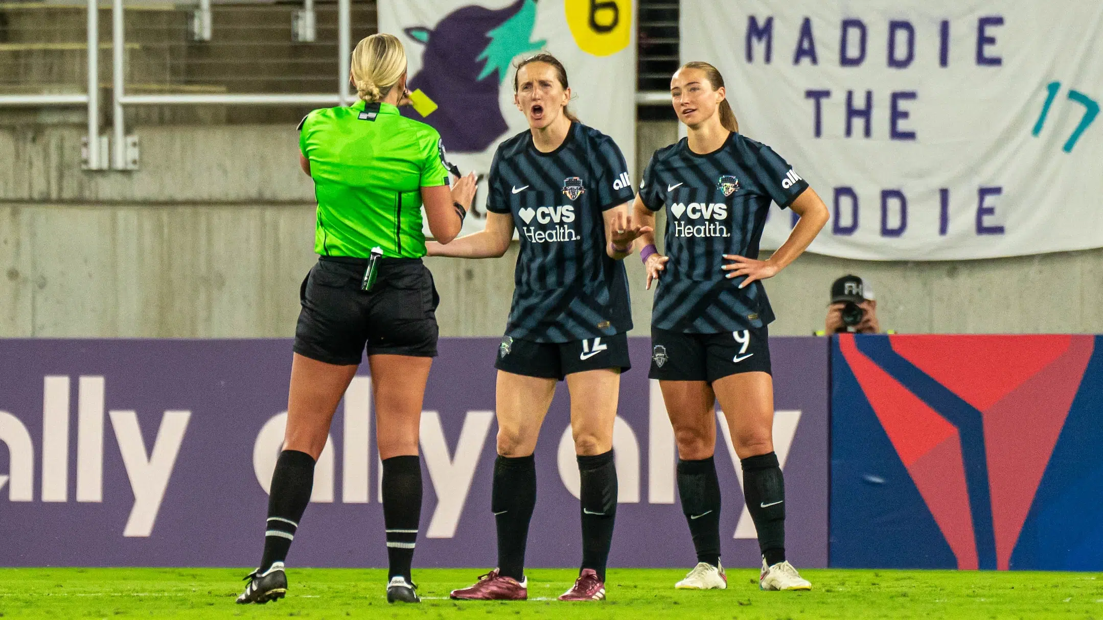 Andi Sullivan exclaims in disgust as a referee in a bright green shirt shows her a yellow card. Tara McKeown stands behind Andi with her hands on her hips.