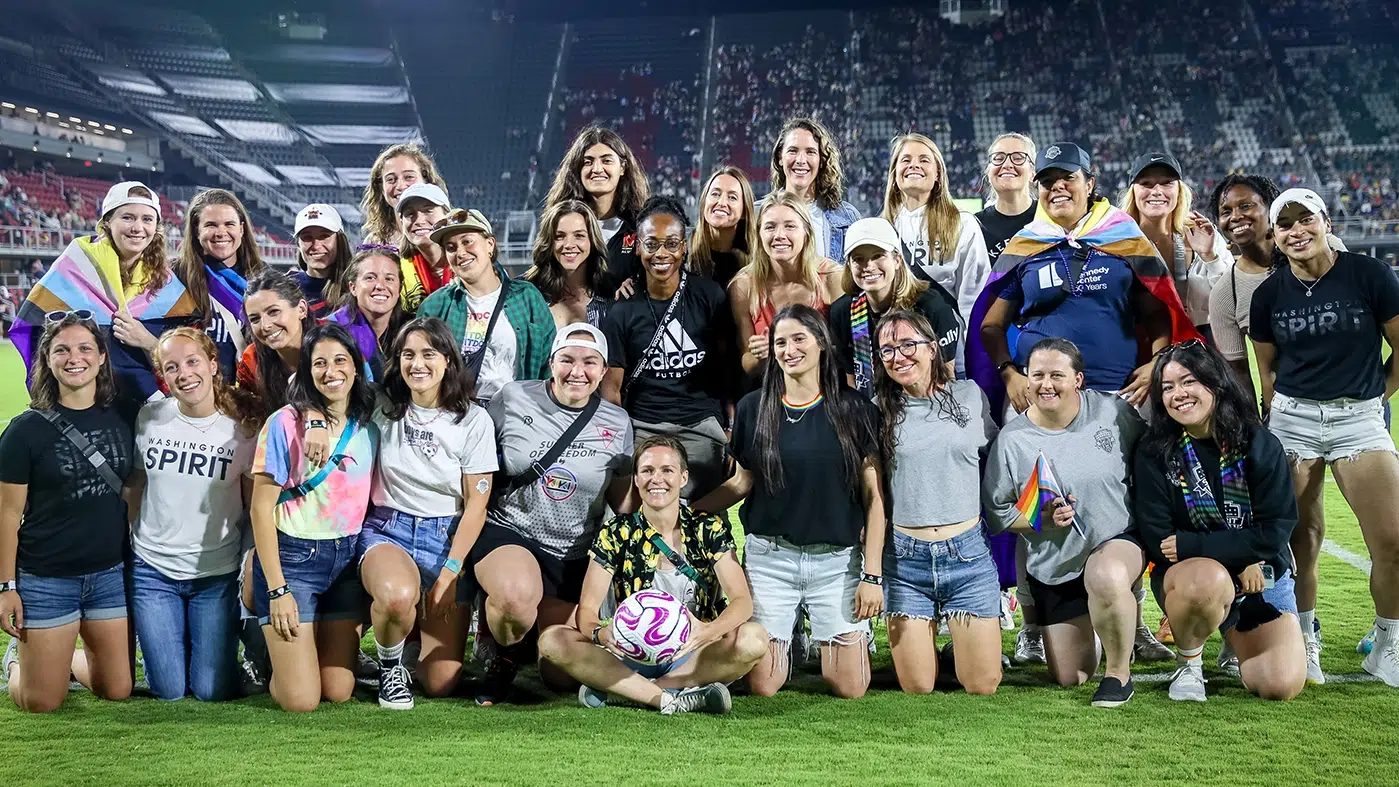 A group of Spirit fans dressed in shorts and t-shirts gathers in three rows for a photo. Their arms are around each other, some wear crossbody bags, some had rainbow flags around their shoulders.