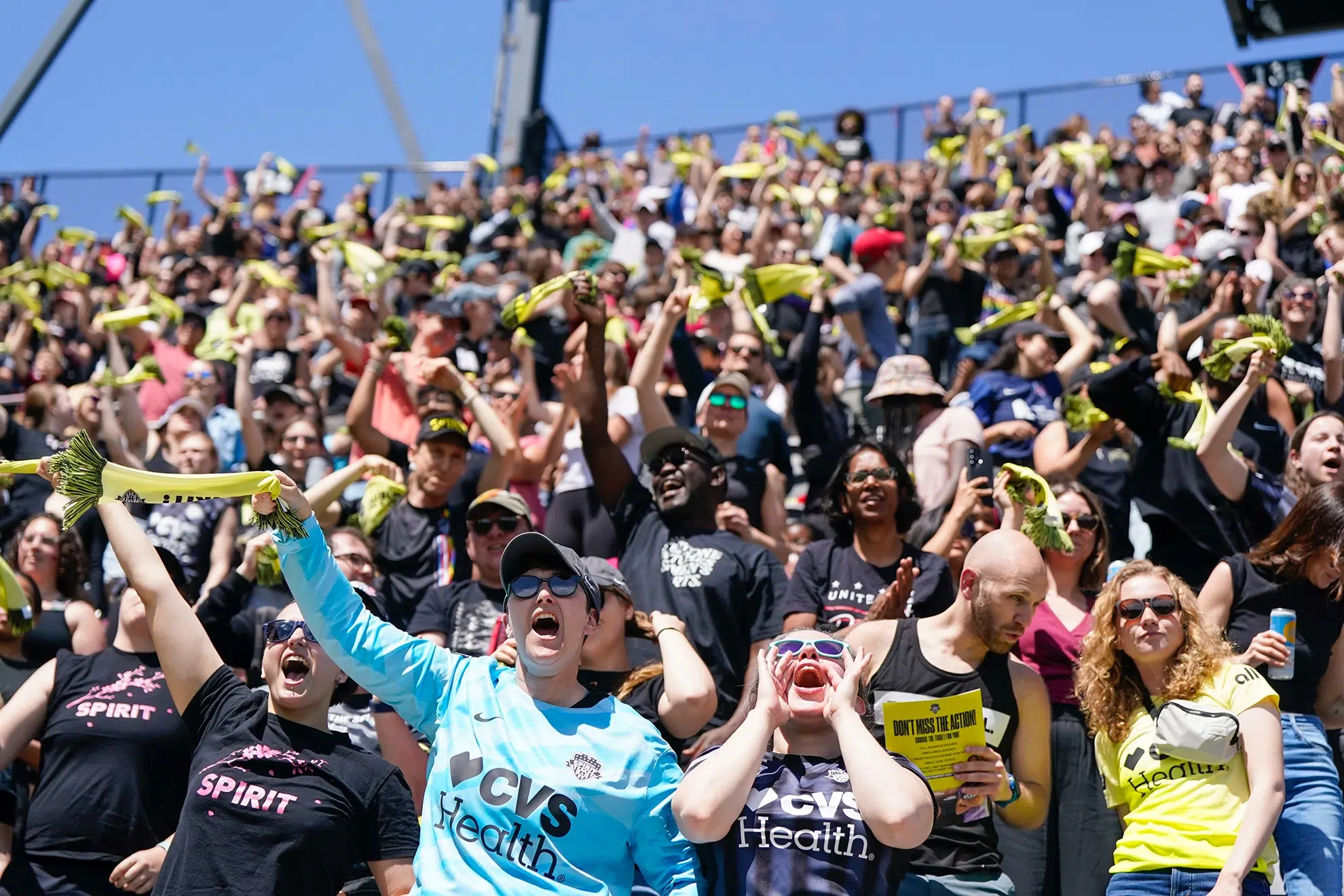 A crowd of fans in various Spirit t-shirts and jerseys wave yellow mini scarves in the air.