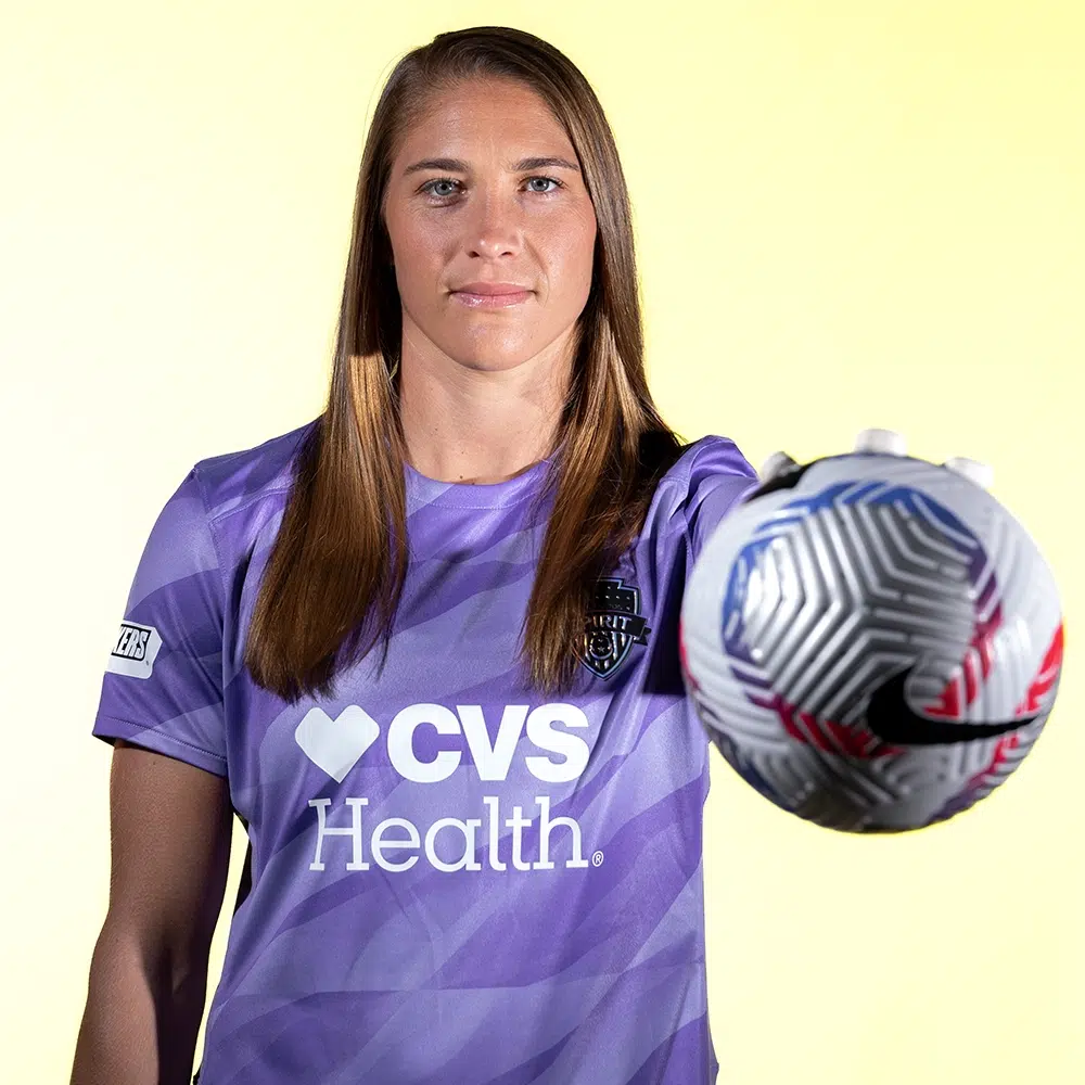 Aubrey Kingsbury in a purple keeper kit holds a ball out with her left hand.