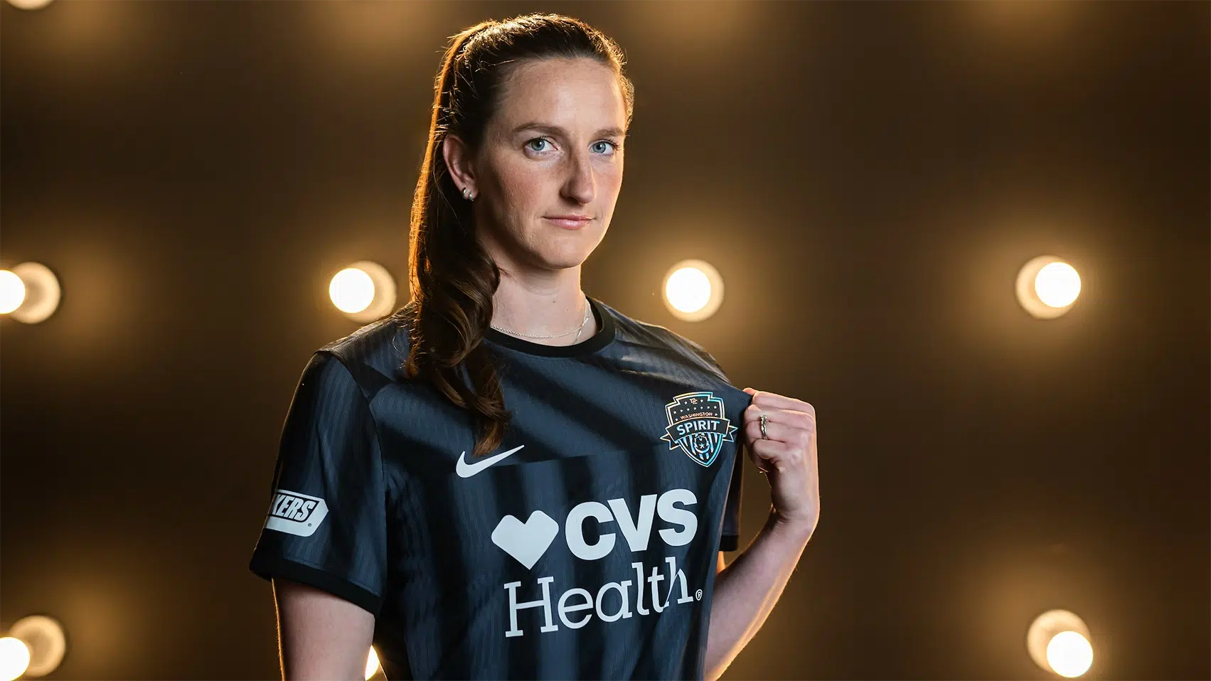 Andi Sullivan holds out the Washington Spirit crest on her black jersey while standing in front of a wall of light bulbs.