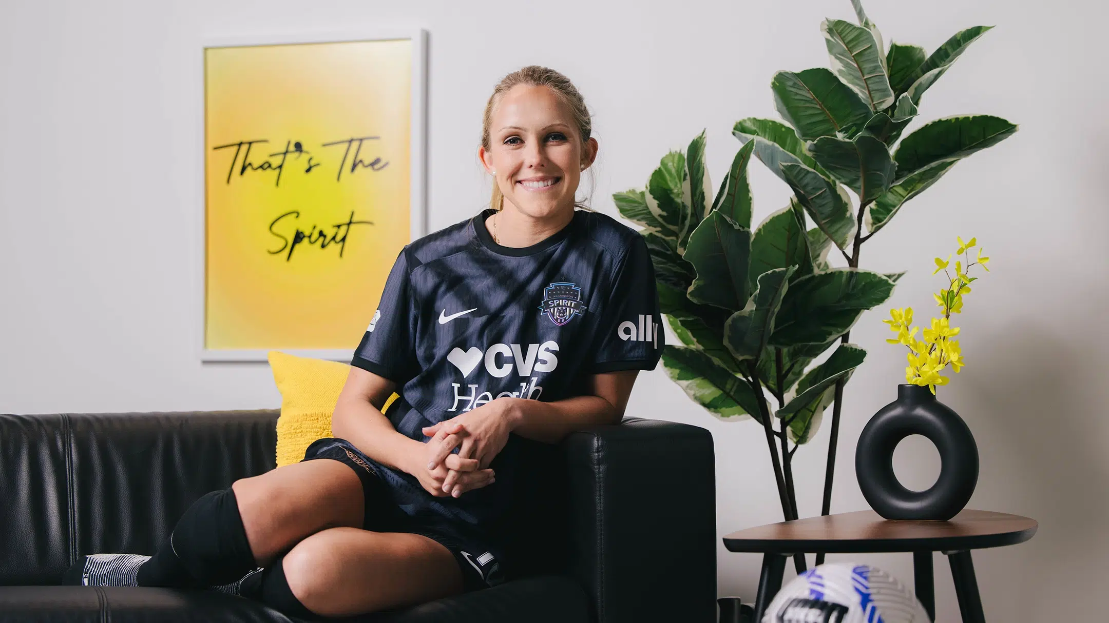 Brittany Ratcliffe sits on a black couch while wearing a black and gray Spirit kit. In the background is a green plant and a yellow, framed posted that reads "That's the Spirit".