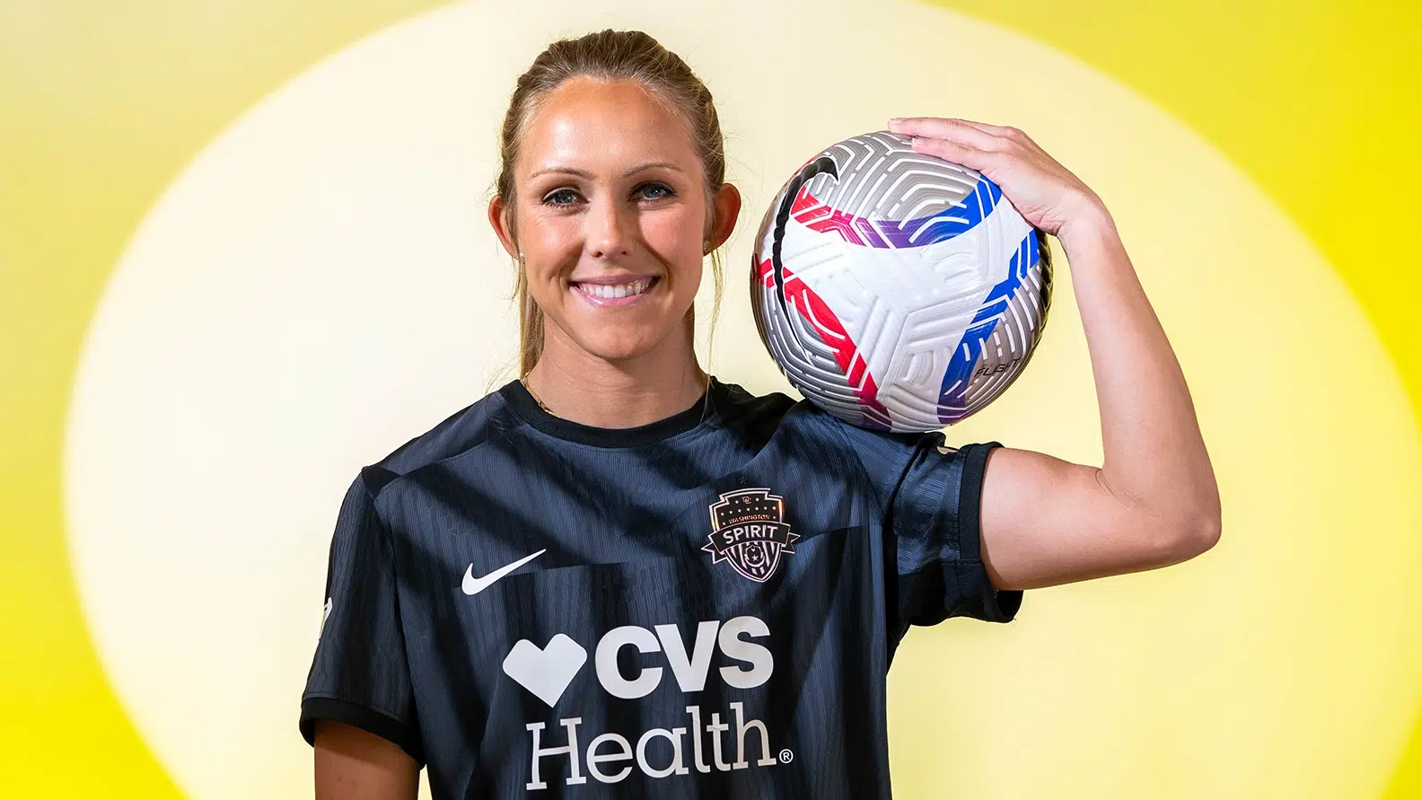 Brittany Ratcliffe smiles and holds a soccer ball on her right shoulder in front of a yellow background.