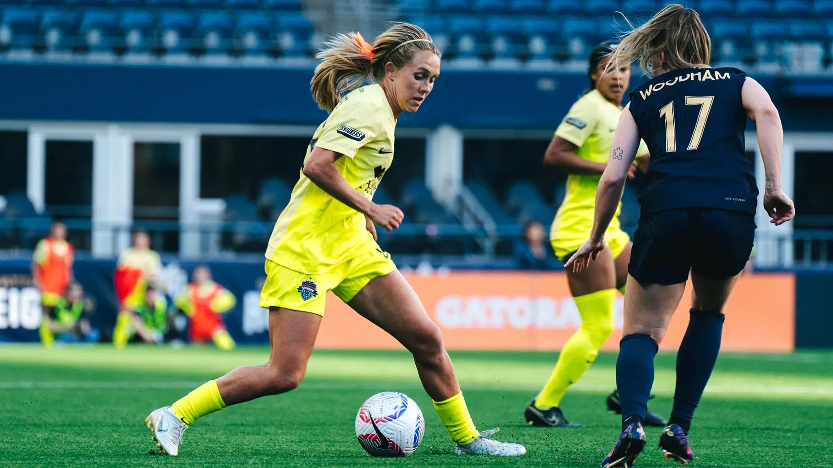 Brittany Ratcliffe dribbles a soccer ball towards a Seattle Reign defender. Brittany is wearing a neon yellow Spirit kit with her hair in a ponytail and orange ribbon.