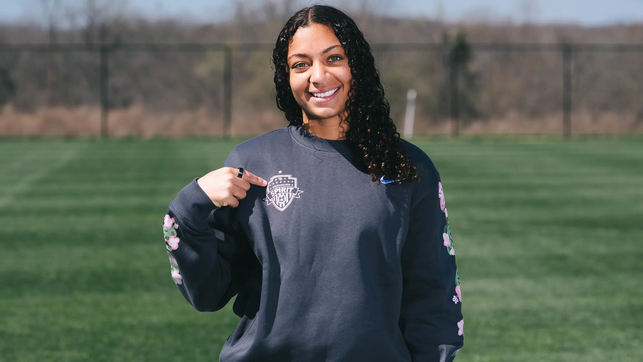 Mikaela Morris stands on a soccer field wearing a dark gray crewneck sweatshirt. She's pointing to the pink Washington Spirit crest on her chest.