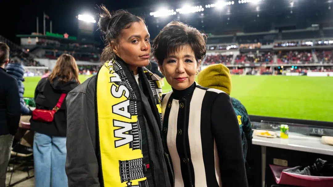 Domo Wells and Michele Kang pose for a photo. The pitch of Audi Field and the stadium lights are visible in the background.