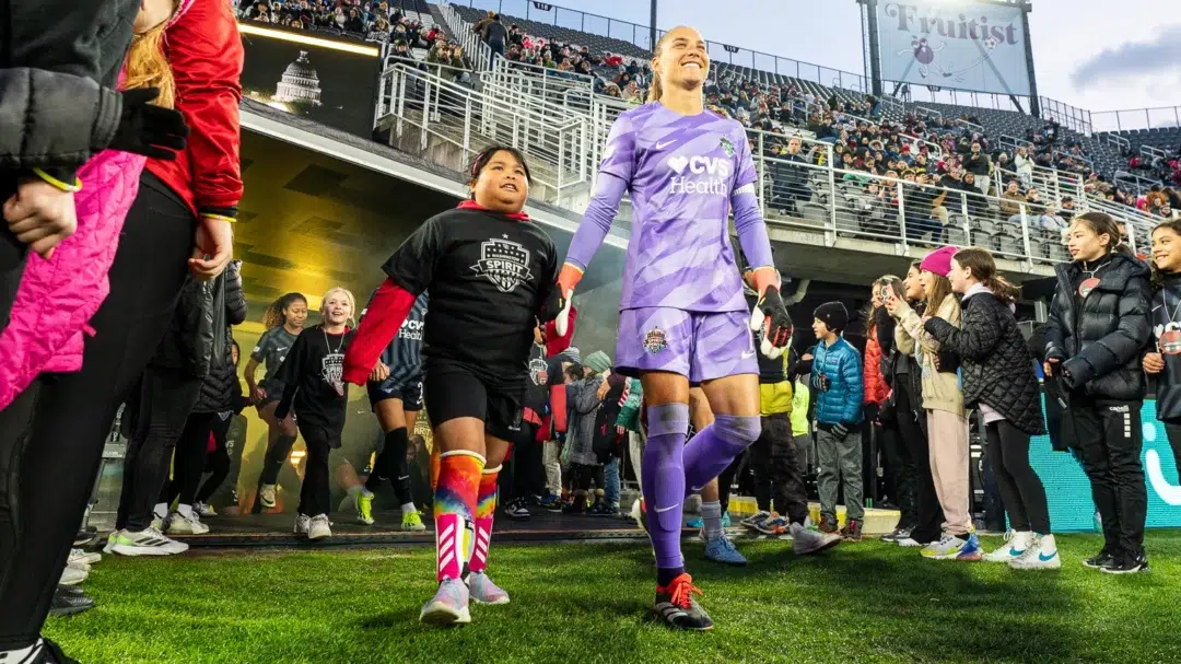Aubrey Kingsbury, wearing a purple goalkeeper kit, walks onto the pitch at Audi Field holding the hand of a young girl in a black Washington Spirit t-shirt.