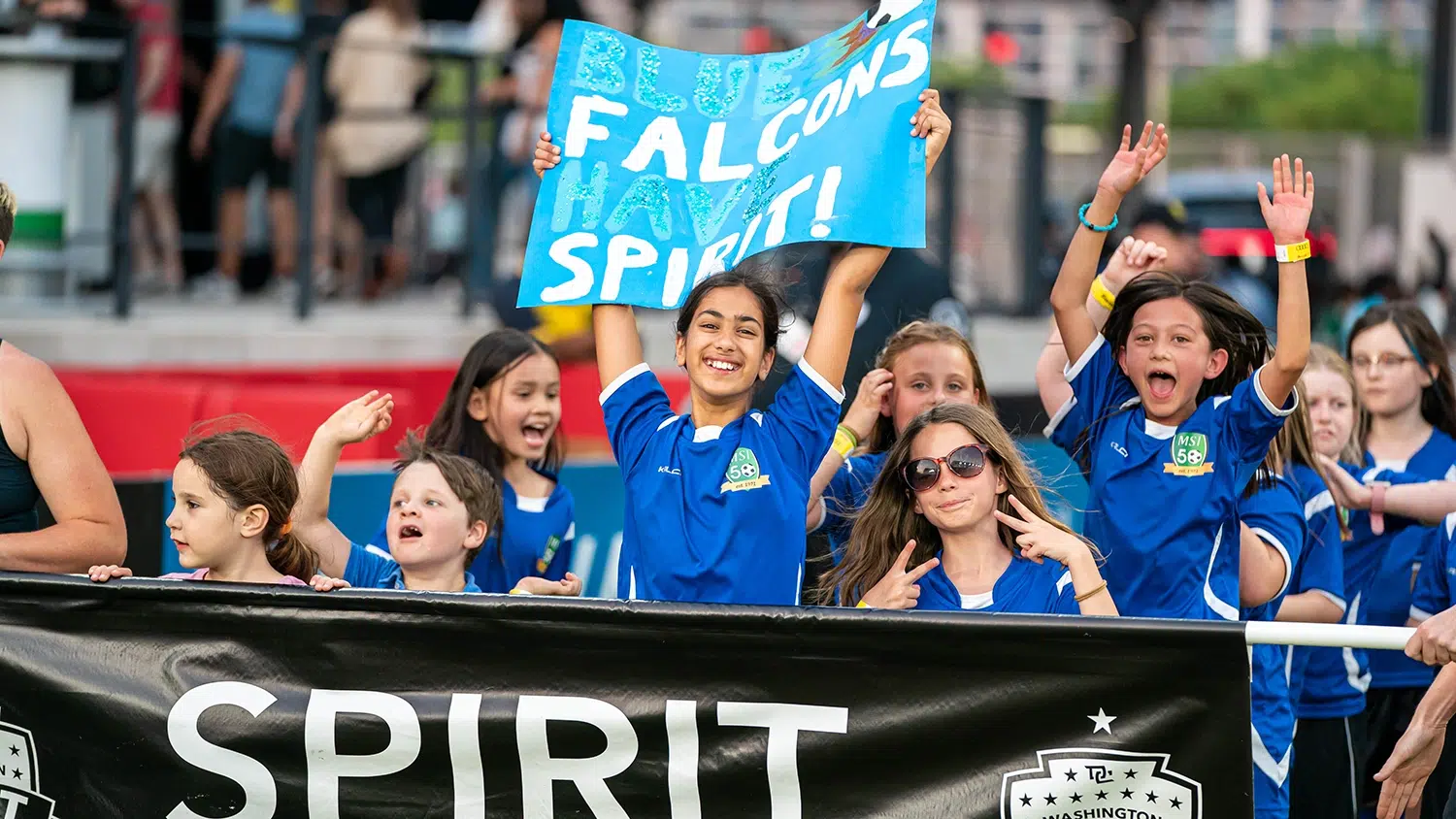 Young girls in matching blue soccer uniforms cheer. The girl in the middle holds up a poster that reads 