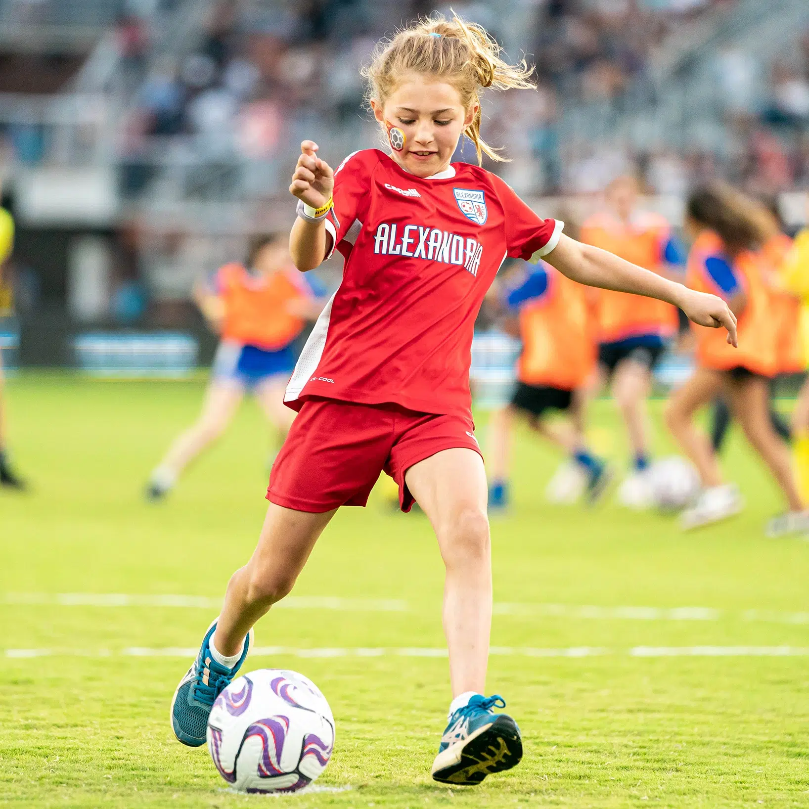 A young girl in a red Alexandria Soccer kit prepares to kick a ball.