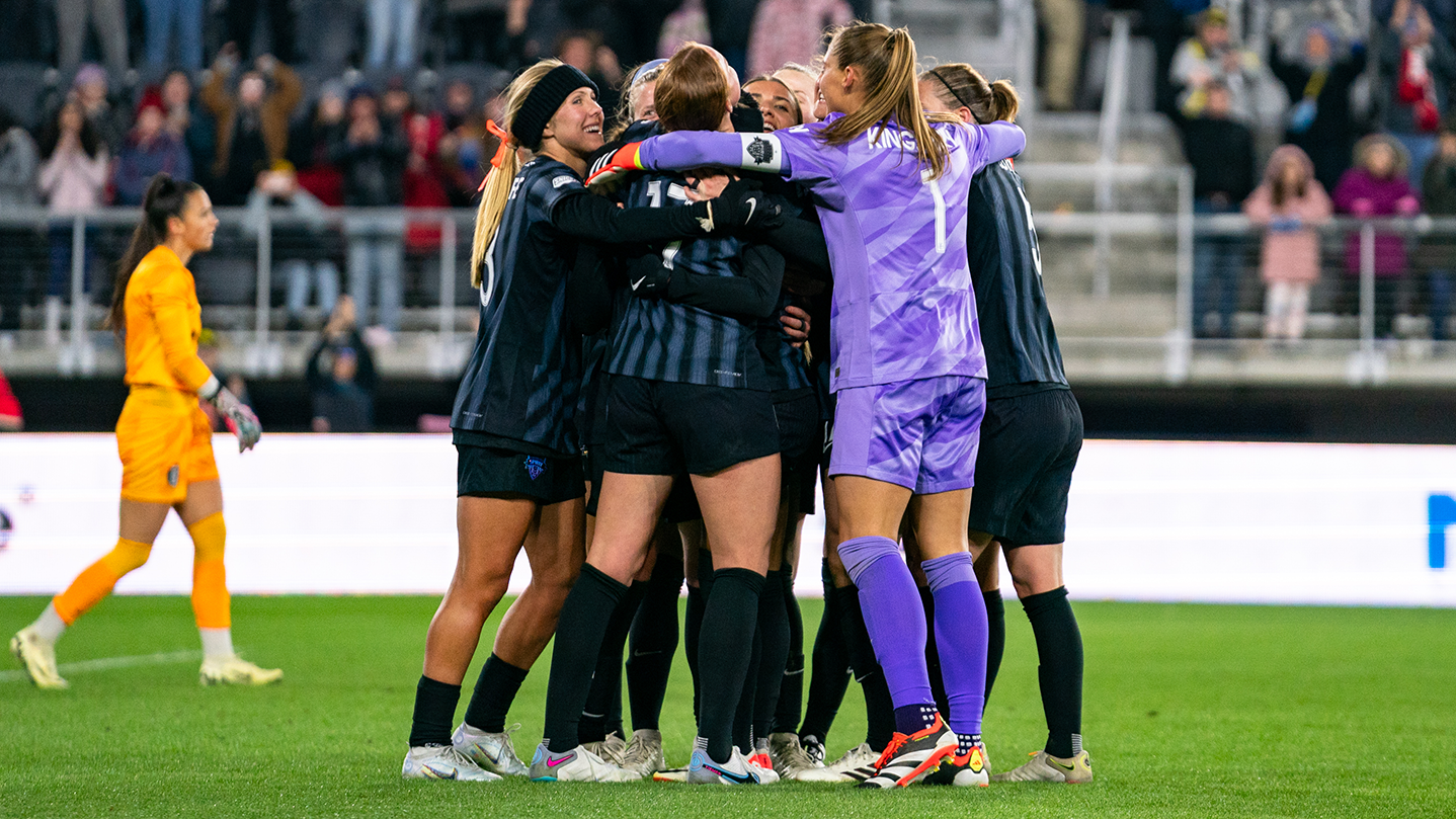 A group of Spirit players in black kits and Aubrey Kingsbury in a purple goalkeeper kit huddle and hug in celebration.