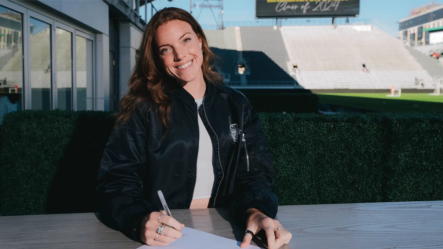 Hal Hershfelt smiles while signing a piece of paper. She's wearing a black satin bomber jacket.