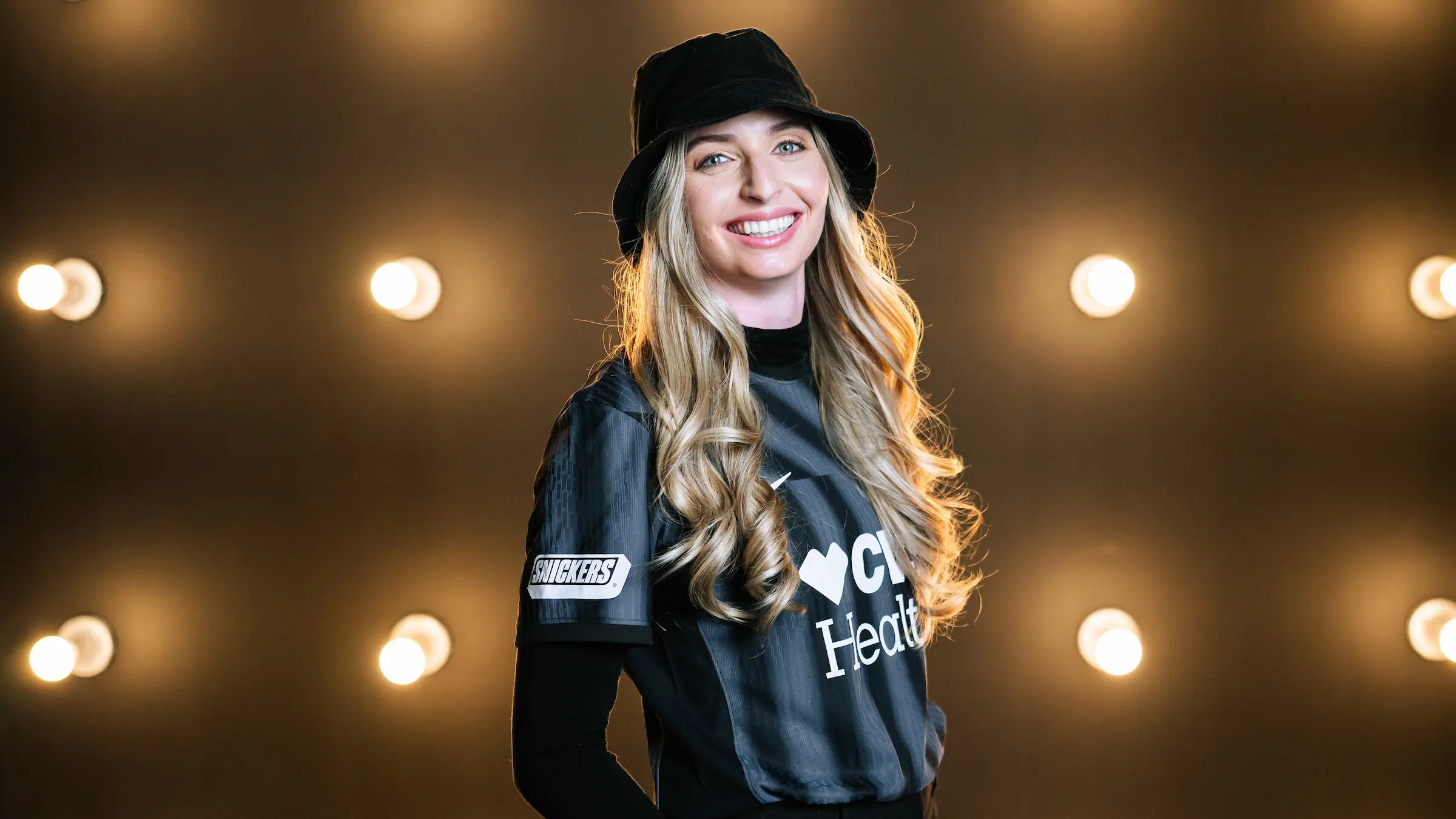 Gabby Carle in a black Spirit jersey and a black bucket hat in front of a wall of light bulbs.