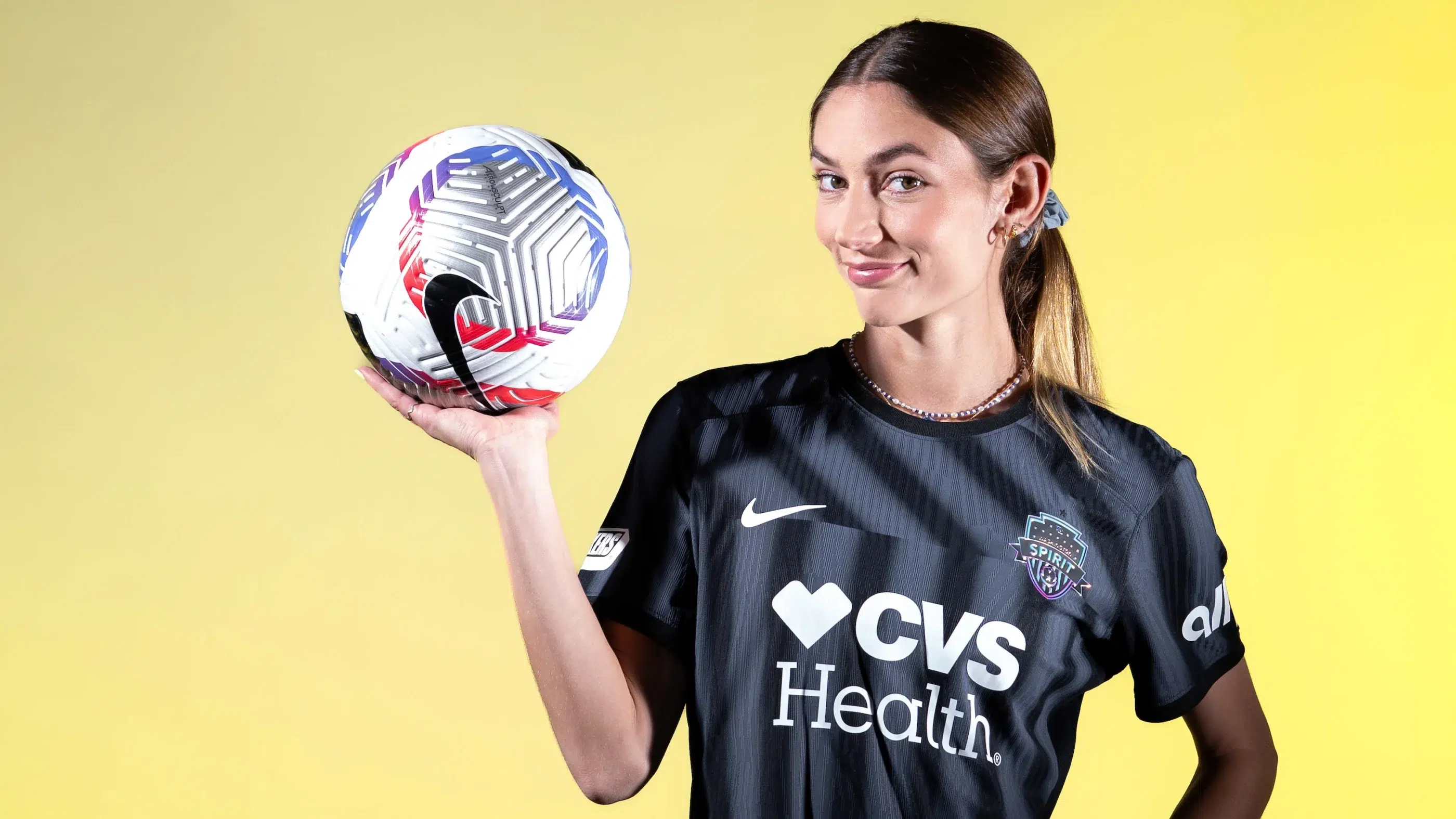 Paige Metayer in a black Spirit jersey holds up a soccer ball in front of a yellow background.