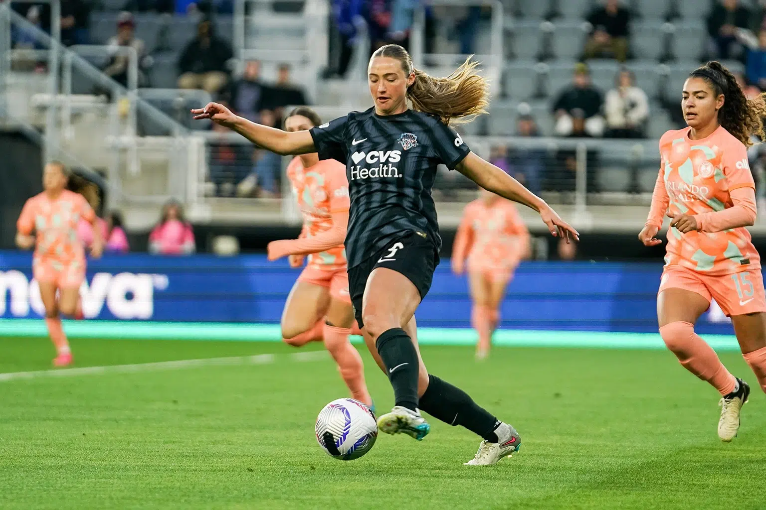 Tara McKeown dribbles a ball while wearing a black kit. Two Orlando defenders in orange kits chase behind.