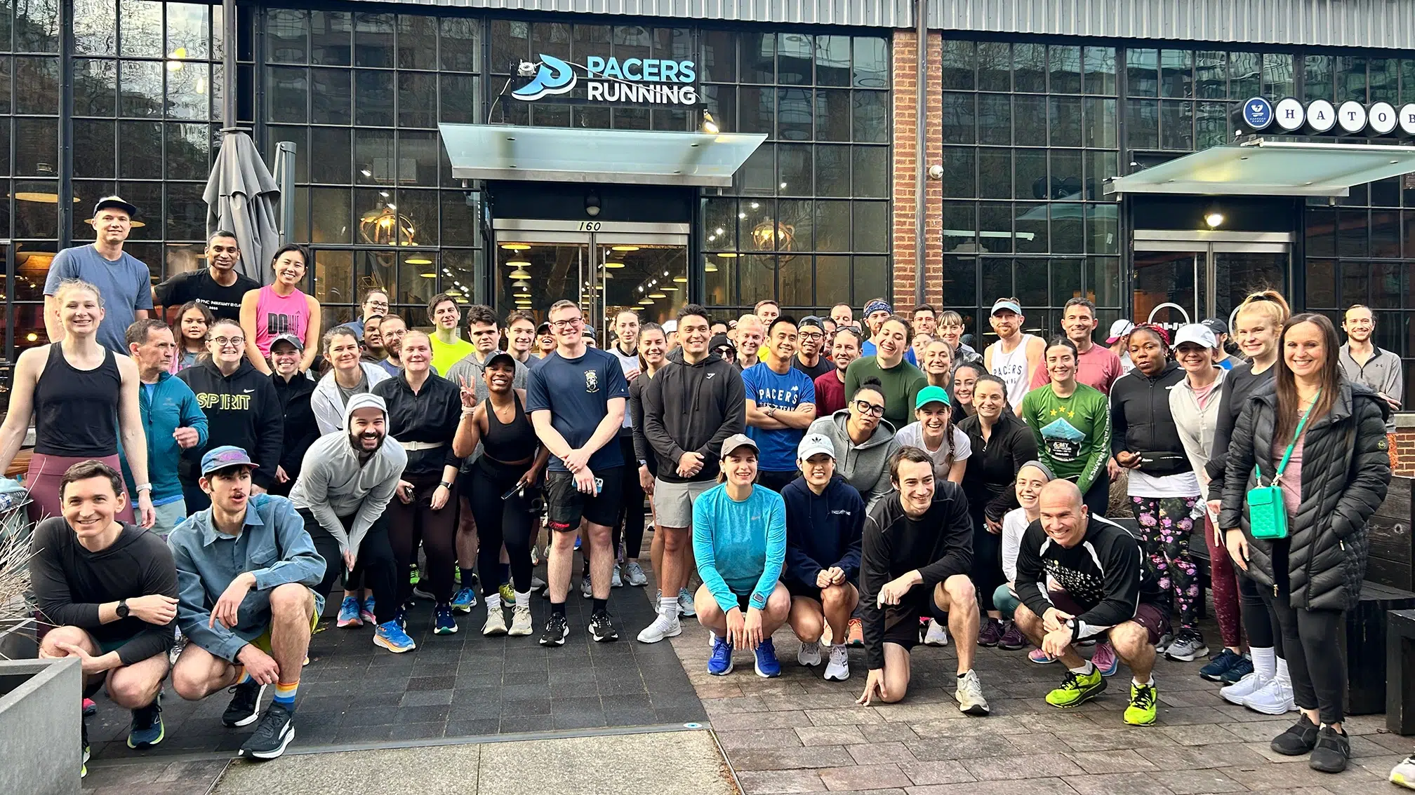 A group of diverse people in running shoes, tank tops, shirts, tights, and shorts pose for a photo in front of a Pacers Running store.