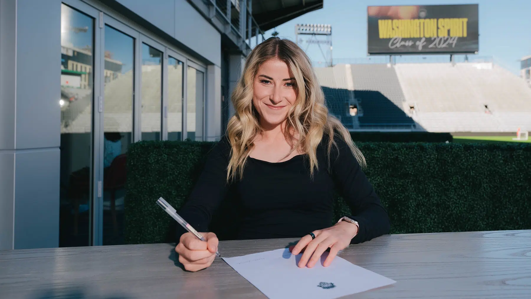 Heather Stainbrook in a black top signs a contract. In the background, Audi Field is seen with "Washington Spirit Class of 2024" on the large stadium screen.