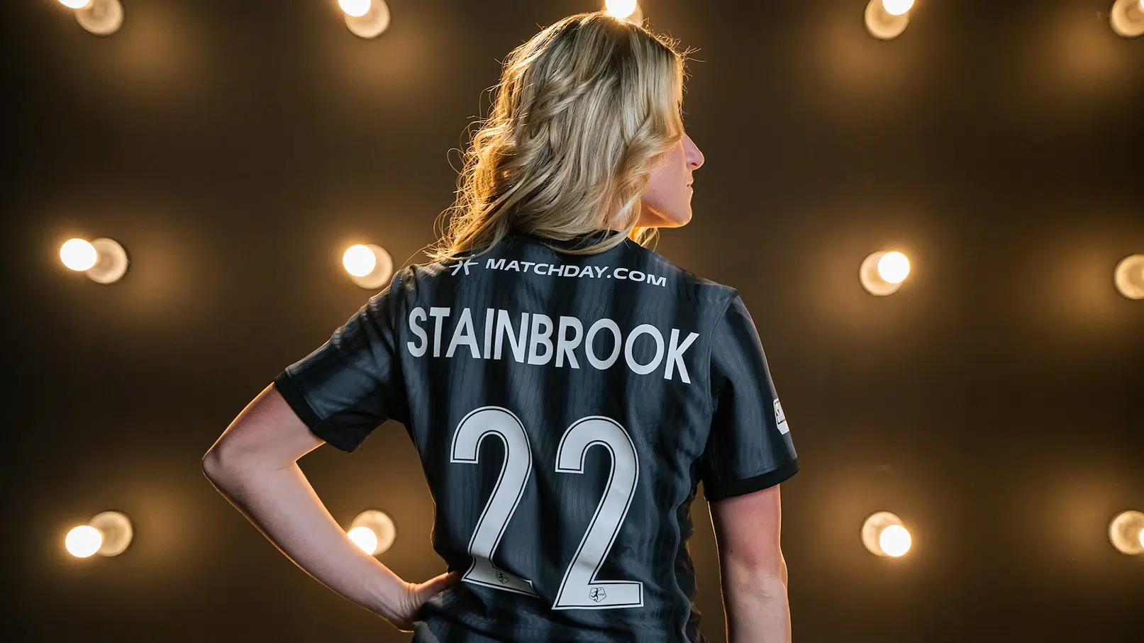 A photo showing the back of Heather Stainbrook's black Spirit jersey. She is standing in front of a wall of lightbulbs and her blonde hair is curled.