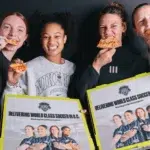 Hal Hershfelt, Croix Bethune, Trinity Rodman, and Andi Sullivan bite into slices of pizza while holding Spirit-branded pizza boxes.