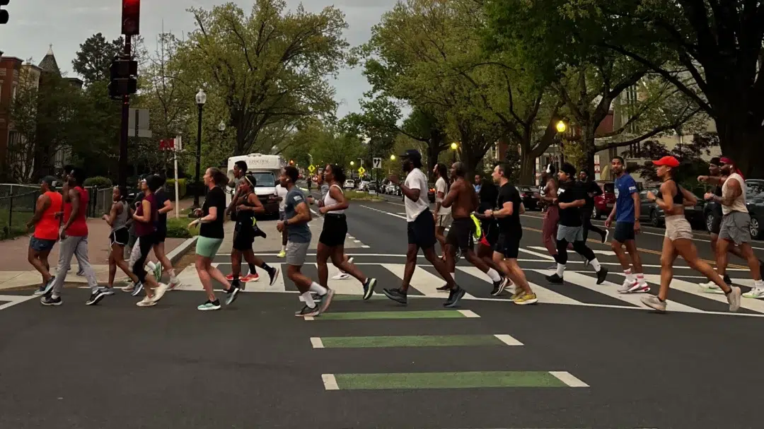 A group of runners crosses the street with trees and rowhouses in the background.