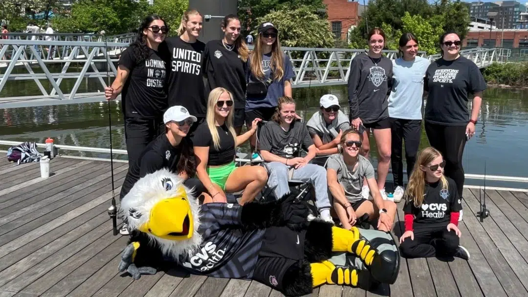 Washington Spirit players in a variety of t-shirts and sweatshirts pose on a dock with fishing rods in hand. A mascot eagle lies in front.