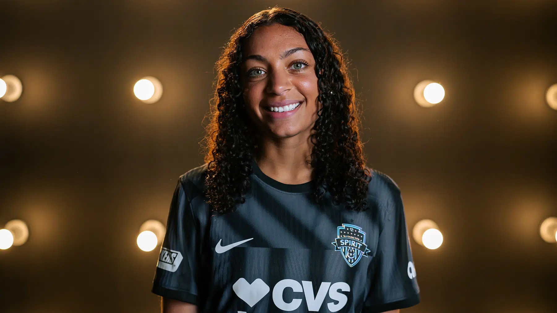 Makenna Morris with her black, curly hair down while wearing a black Spirit jersey and standing in front of a wall of lightbulbs.