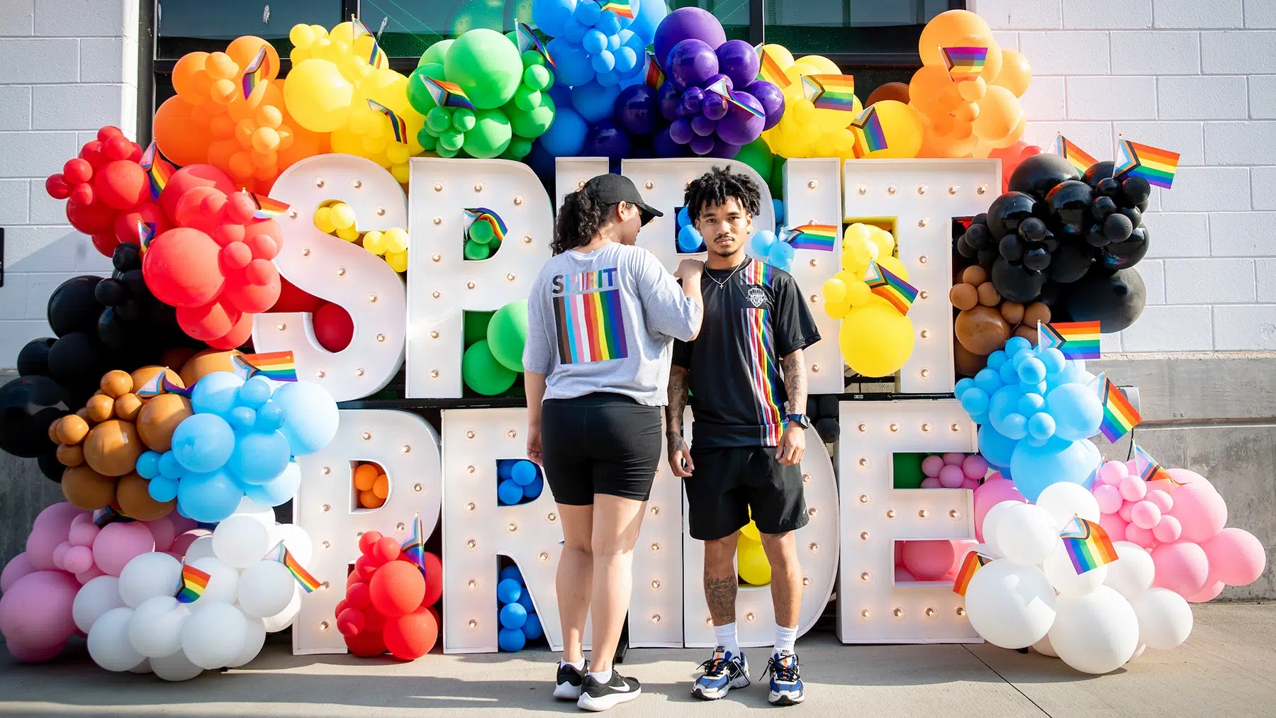 Two people stand in front of marquee letter spelling out "SPIRIT PRIDE" with colorful balloons.