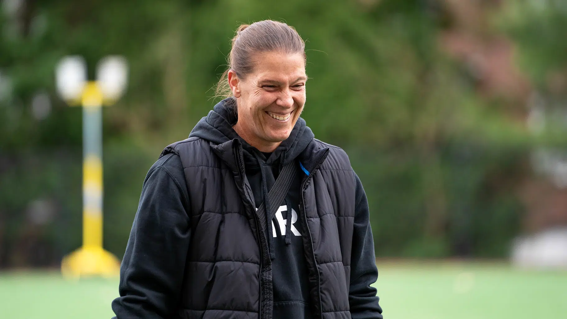 Nicole Barnhart in a black sweatshirt and jacket smiles on a soccer field.
