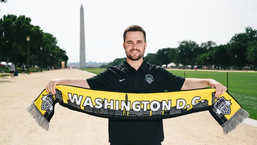 Jonatan Giraldez standing in front of the Washington Monument holding a Spirit scarf.
